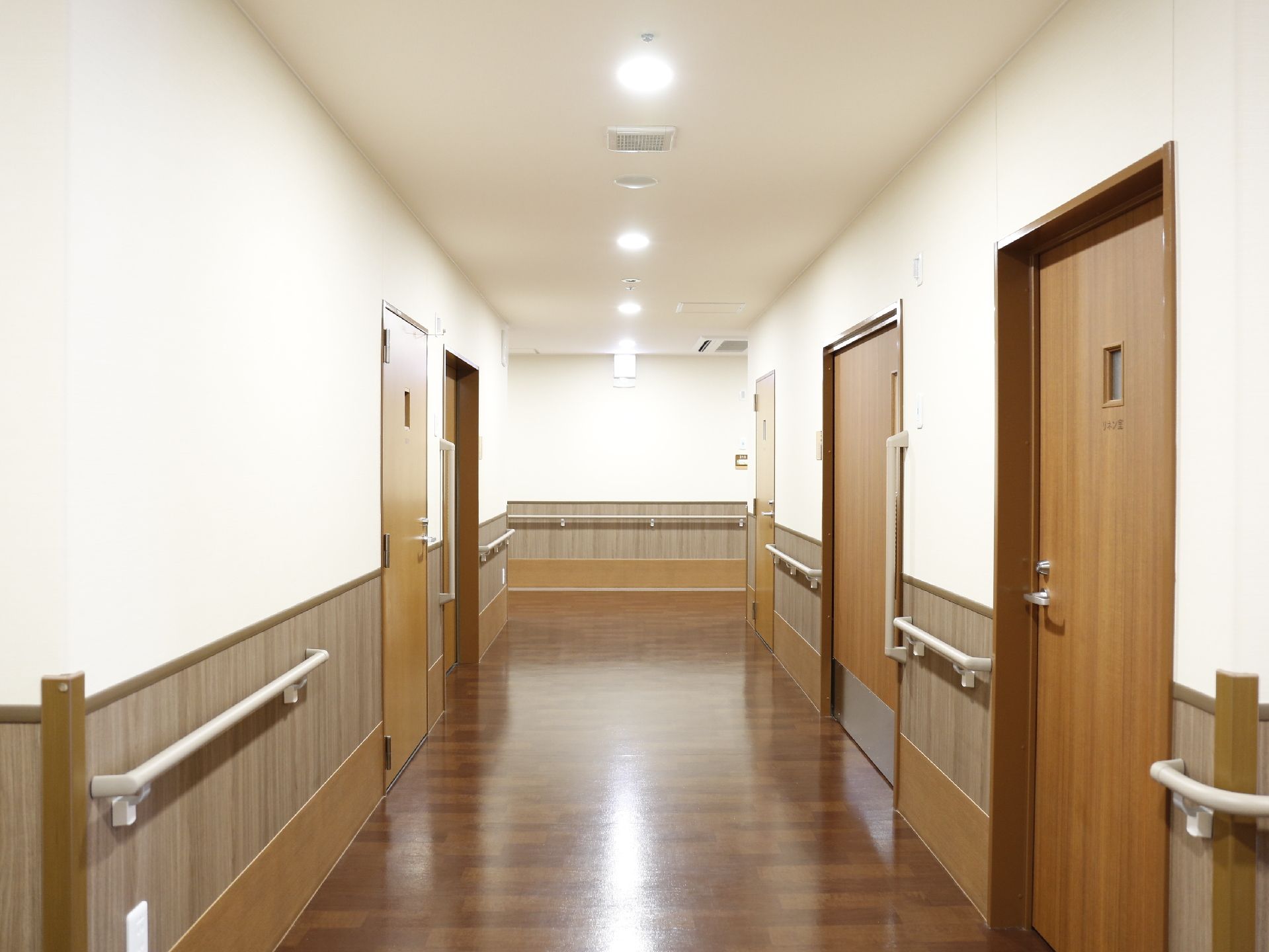 通路のすべての壁に横向きの手すりと引き戸の部屋の取っ手側に縦向きの手すりがある。天井にはほぼ等間隔のライトがついている。