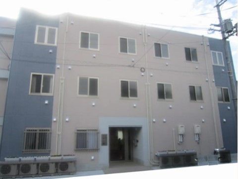 ベージュとグレー、２色の外壁になっている落ち着いた雰囲気の建物である。玄関があるスペースには屋根があり、雨の日も安心である。