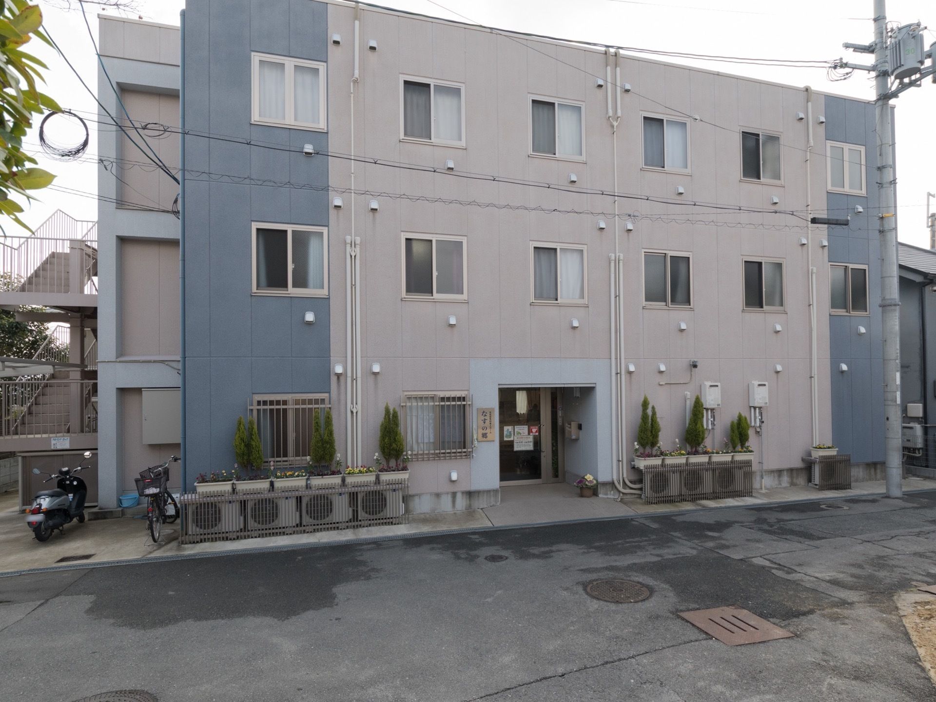 グレーとベージュのツートンカラーの外壁がおしゃれな、３階建てのアパートのような外観である。入り口には施設名を掲げた看板もある。