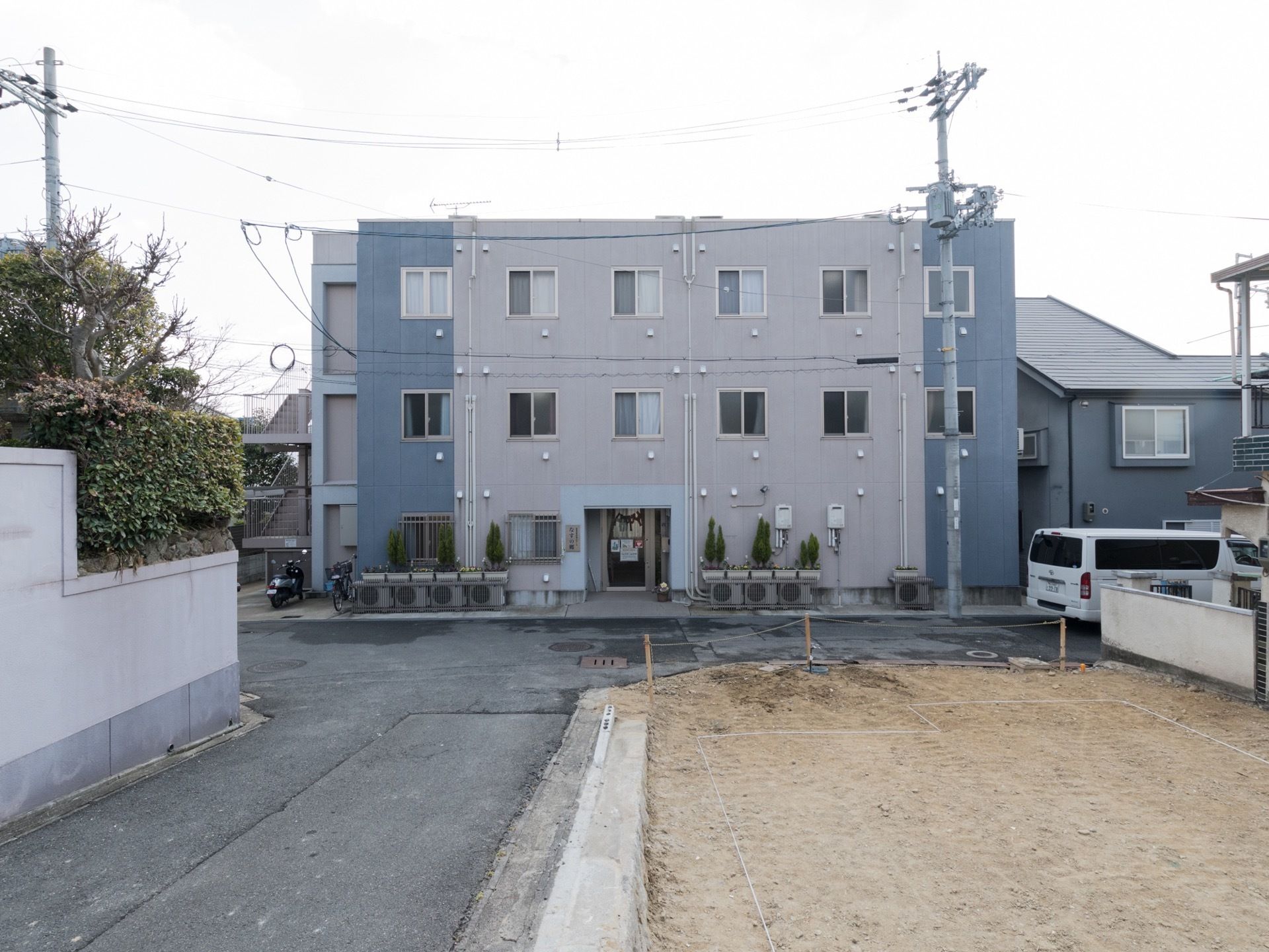 閑静な住宅街の中にある施設は、グレーとベージュのツートンカラーの外壁が特徴的である。T字路の目の前に建っている。