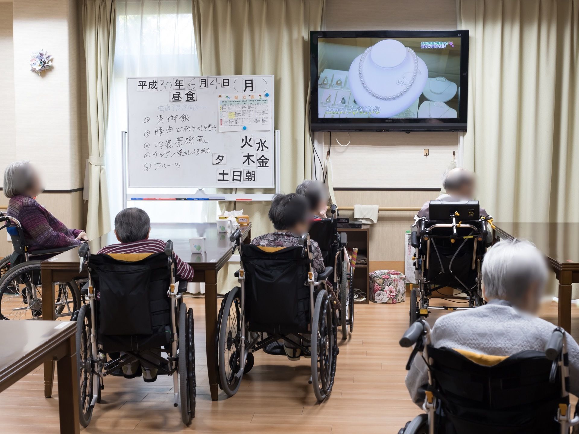 共有スペースにはテレビがあり、複数の入居者様が一緒にテレビ番組を楽しんでいる。テレビに向かって左手にはホワイトボードが設置されている。
