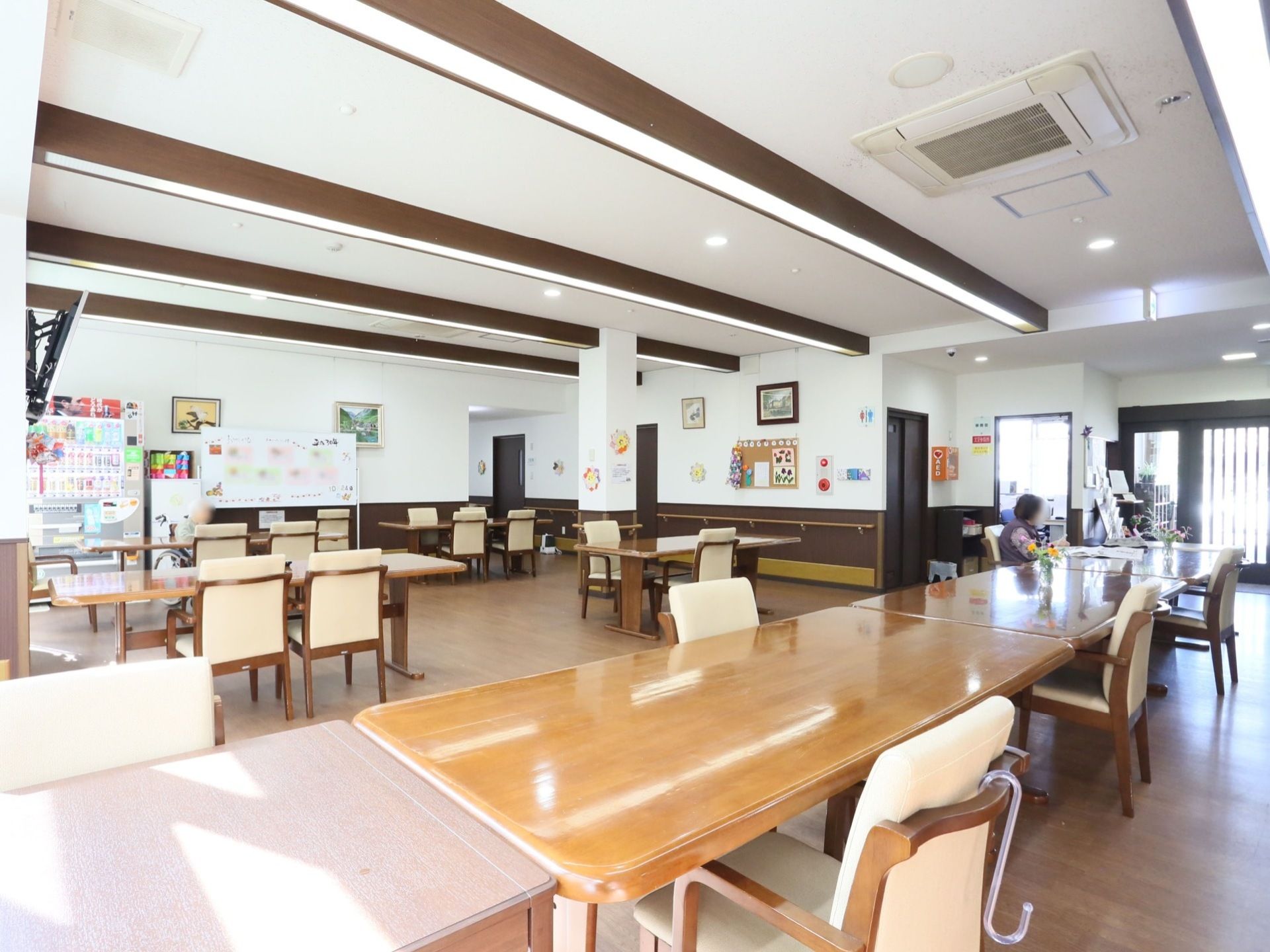 長方形の木製テーブルと椅子のセットが並べて置かれている。席に座り過ごされている入居者様がいらっしゃる。