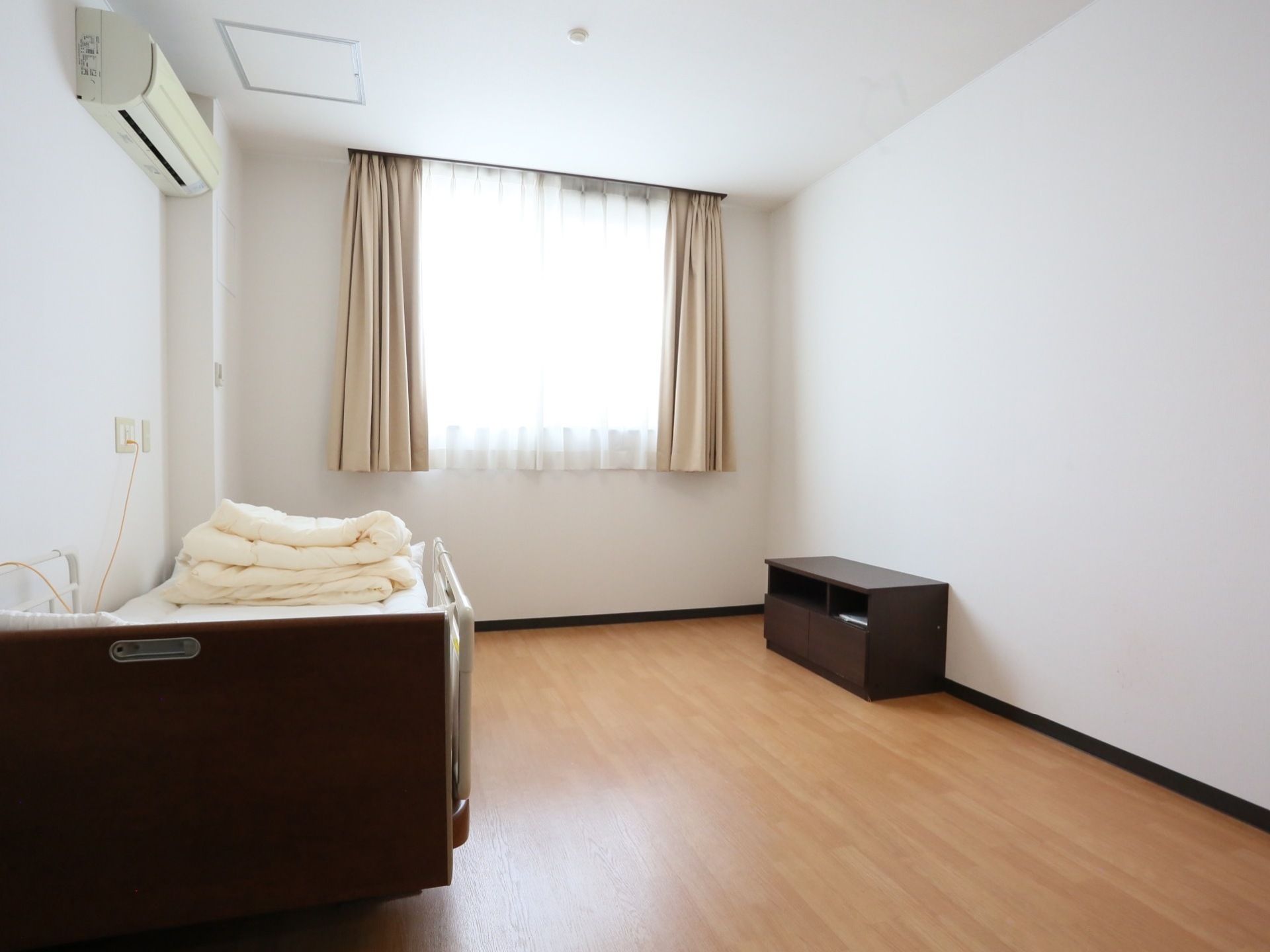 居室には窓があり、カーテンが掛けられている。天井近くにエアコンが取りつけられ、その下には介護用ベッドが置かれている。