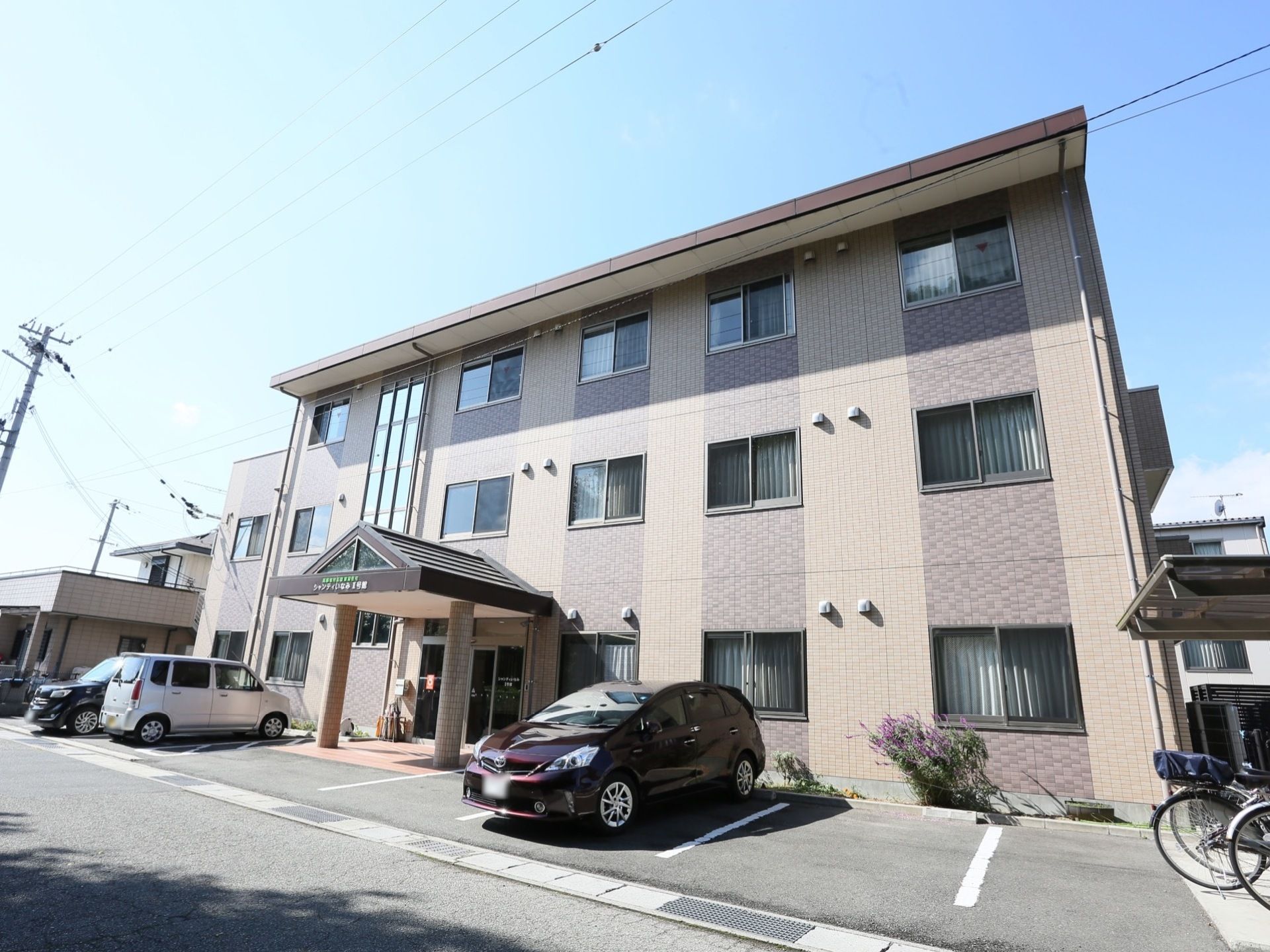 ２色の茶色系に塗り分けられた３階建ての建物である。施設の前は駐車スペースになっており、車が２台停められている。