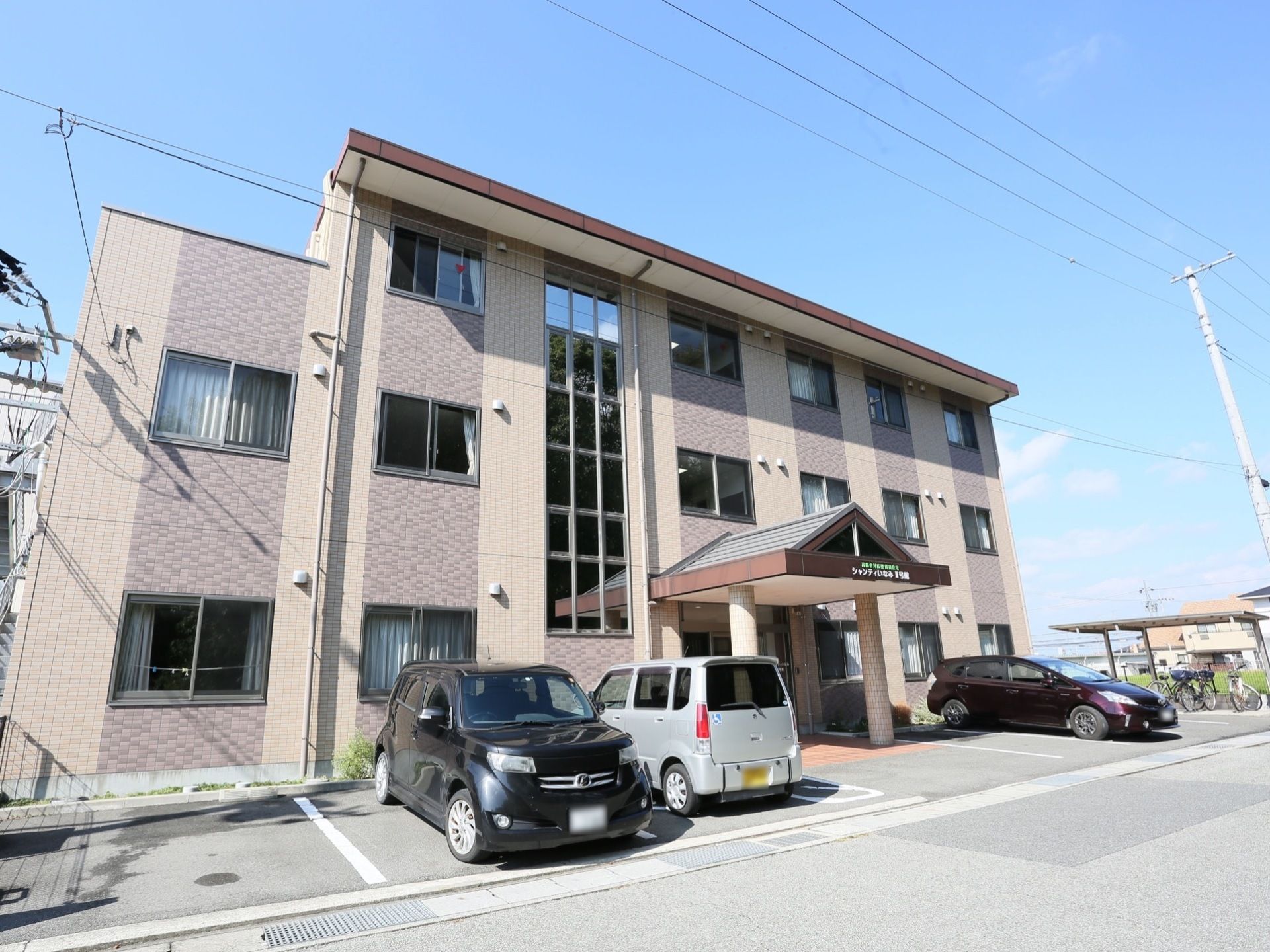 ３階建ての建物で、外壁はブラウン系の２色の縦縞に塗られている。施設前には駐車場があり、車が停まっている。奥に自転車置き場もある。