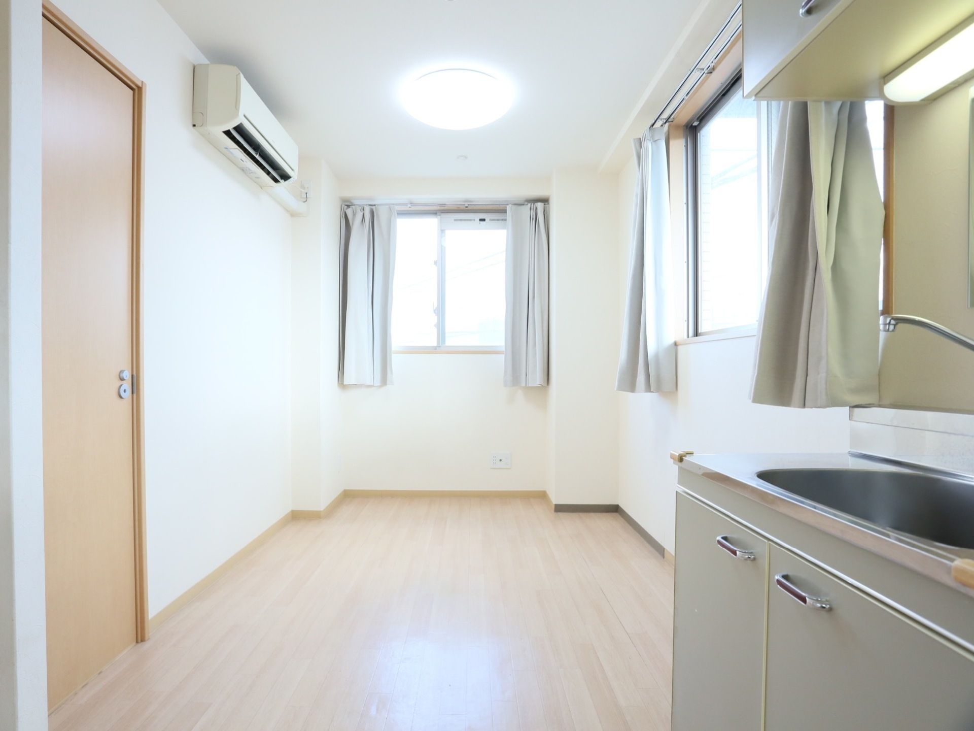 明るいカラーの床材を使用した、フローリングの居室である。窓が２枚備えつけられているほか、エアコンやキッチンもある。