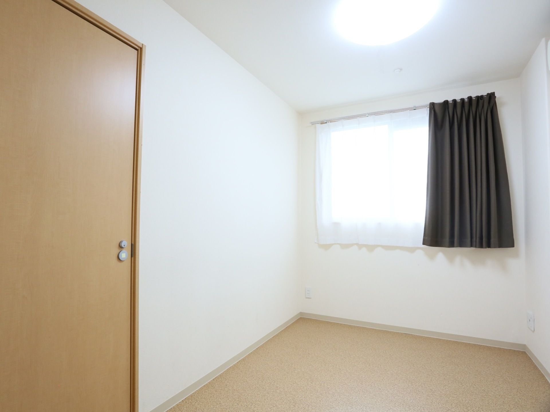 あたたかみのあるフローリング、白い壁に施工された居室である。窓にはレースカーテン、遮光性のあるダーク調のカーテンも備えつけられている。