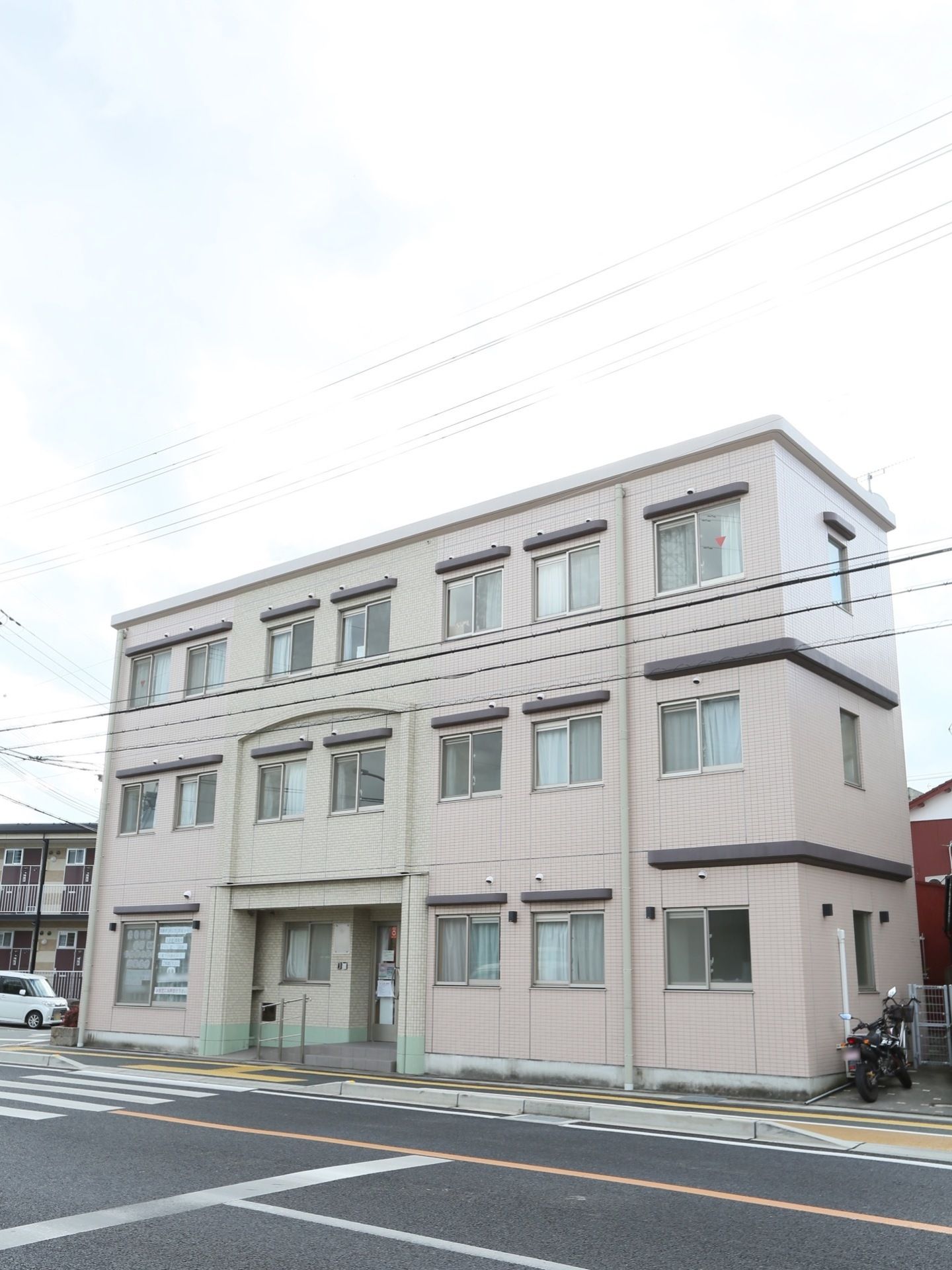 道路に面した場所に立地する、３階建ての施設である。落ち着いた色調の外観。エントランスを出ると、横断歩道がある。