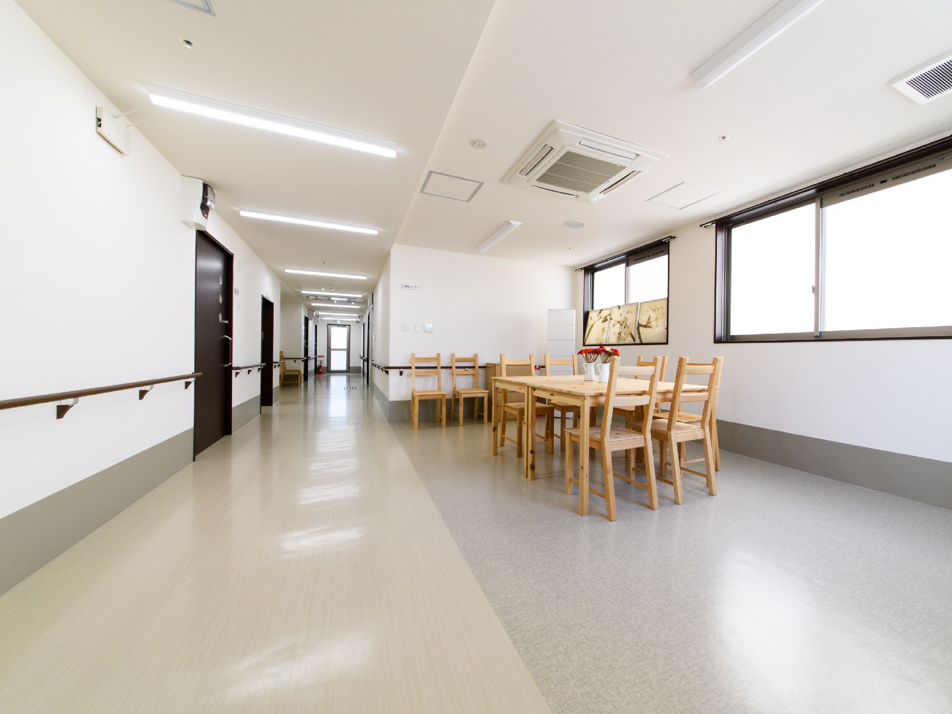 白い壁とグレーの床が明るく開放的な雰囲気の廊下。木製のテーブルと椅子が置かれた共有スペースが設けられている。