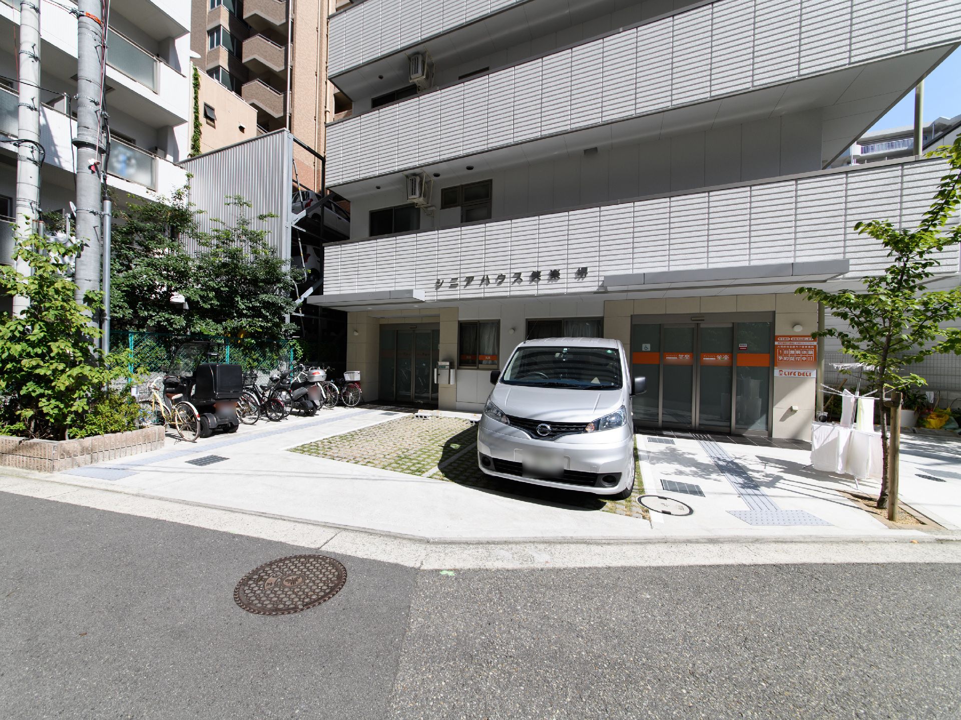 明るい道路に面したグレー系の外壁が落ち着いた雰囲気の施設の外観。入り口の前はコンクリ―トで整備され、駐車スペースが設けられている。