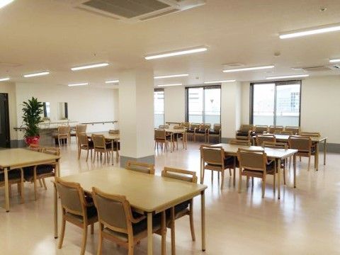 明るい色の床にテーブルと椅子が並んでいる、窓の多い食堂