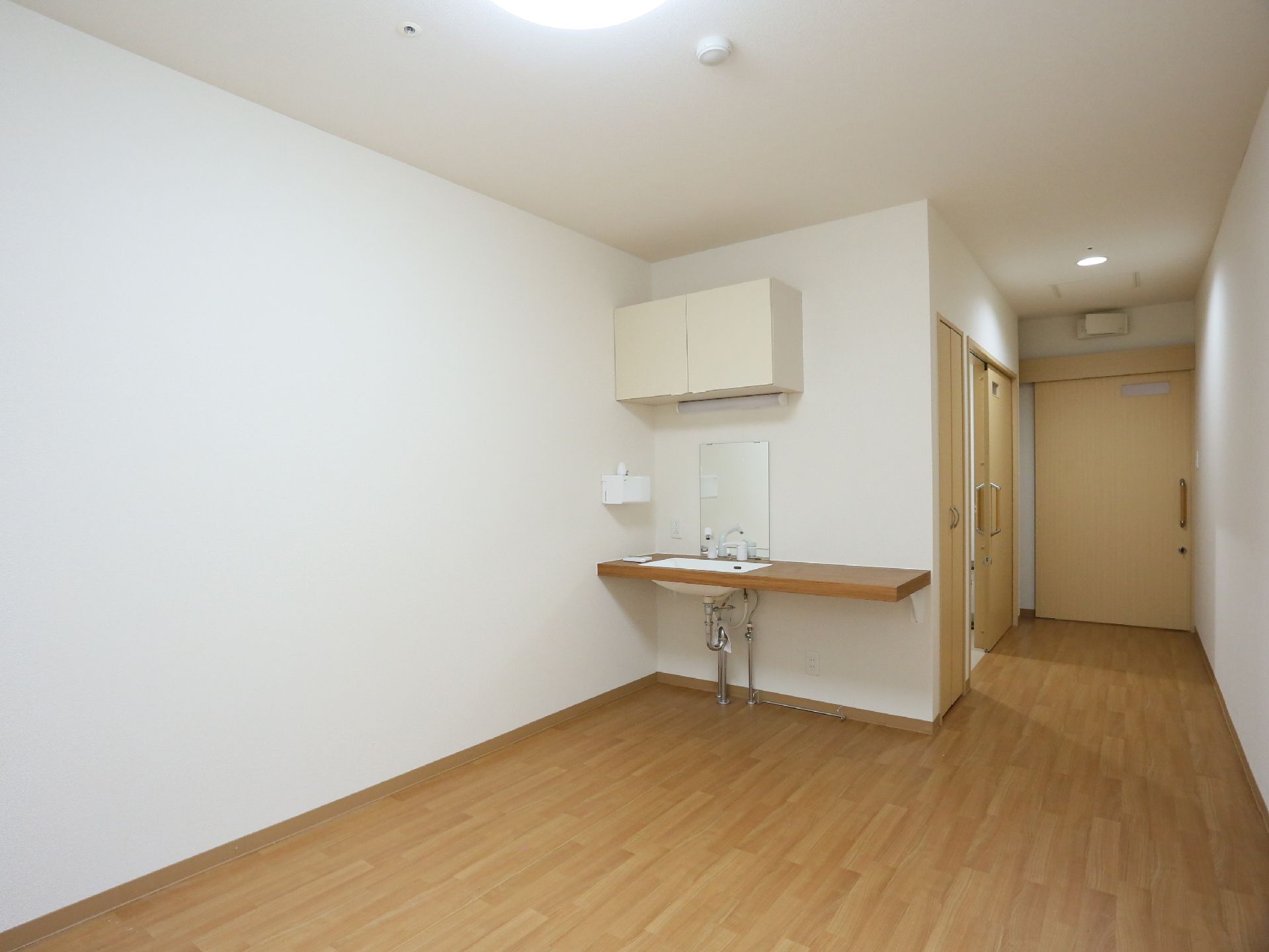入口ドアが奥に見えている。部屋へ入るとトイレと収納のドアがある。居住スペースに洗面台があり、その上に収納棚がある。