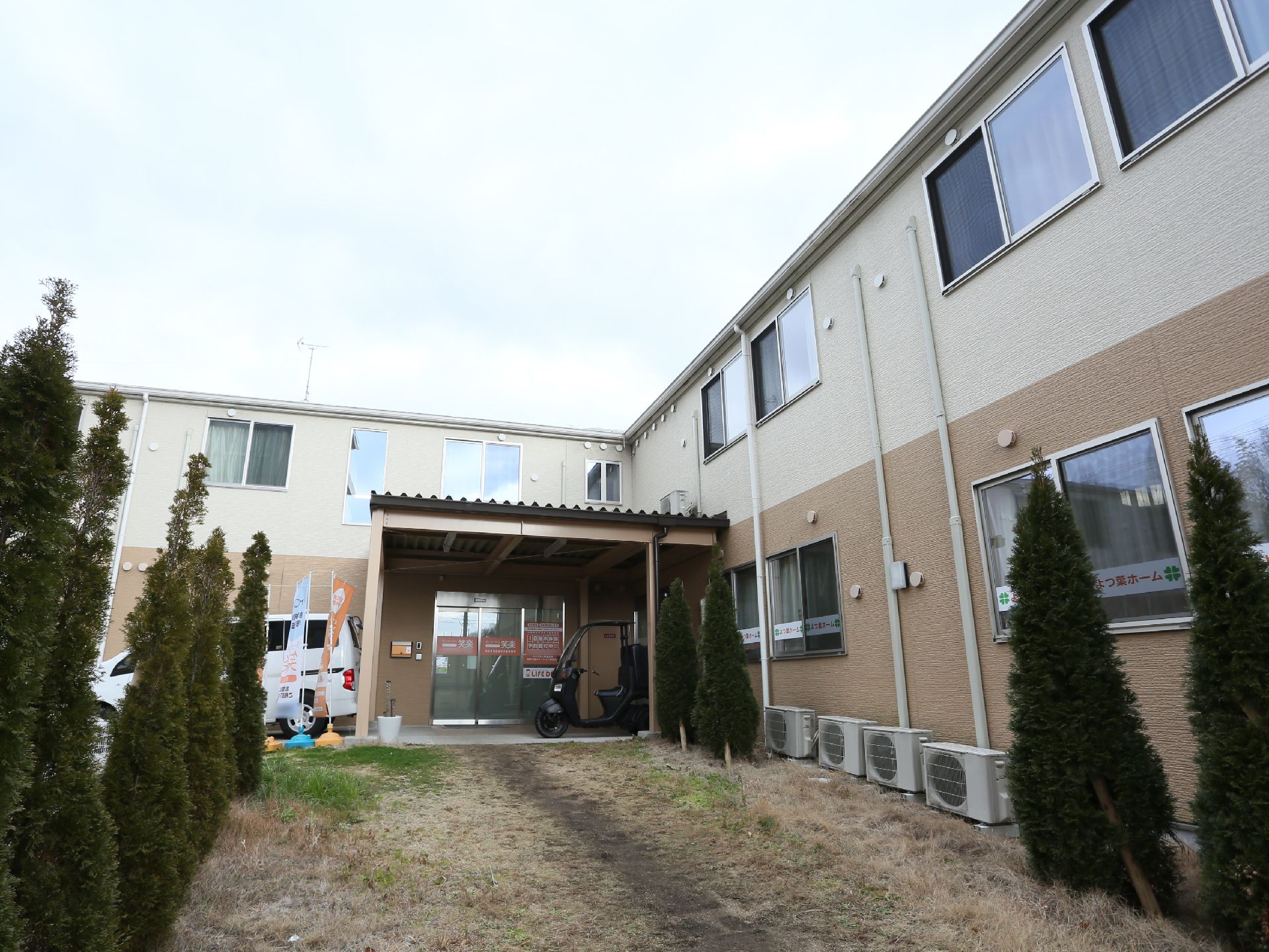 ベージュと茶色のツートンに塗られた外観の施設で２階建てである。玄関部分は柱に支えられた屋根がせり出している。