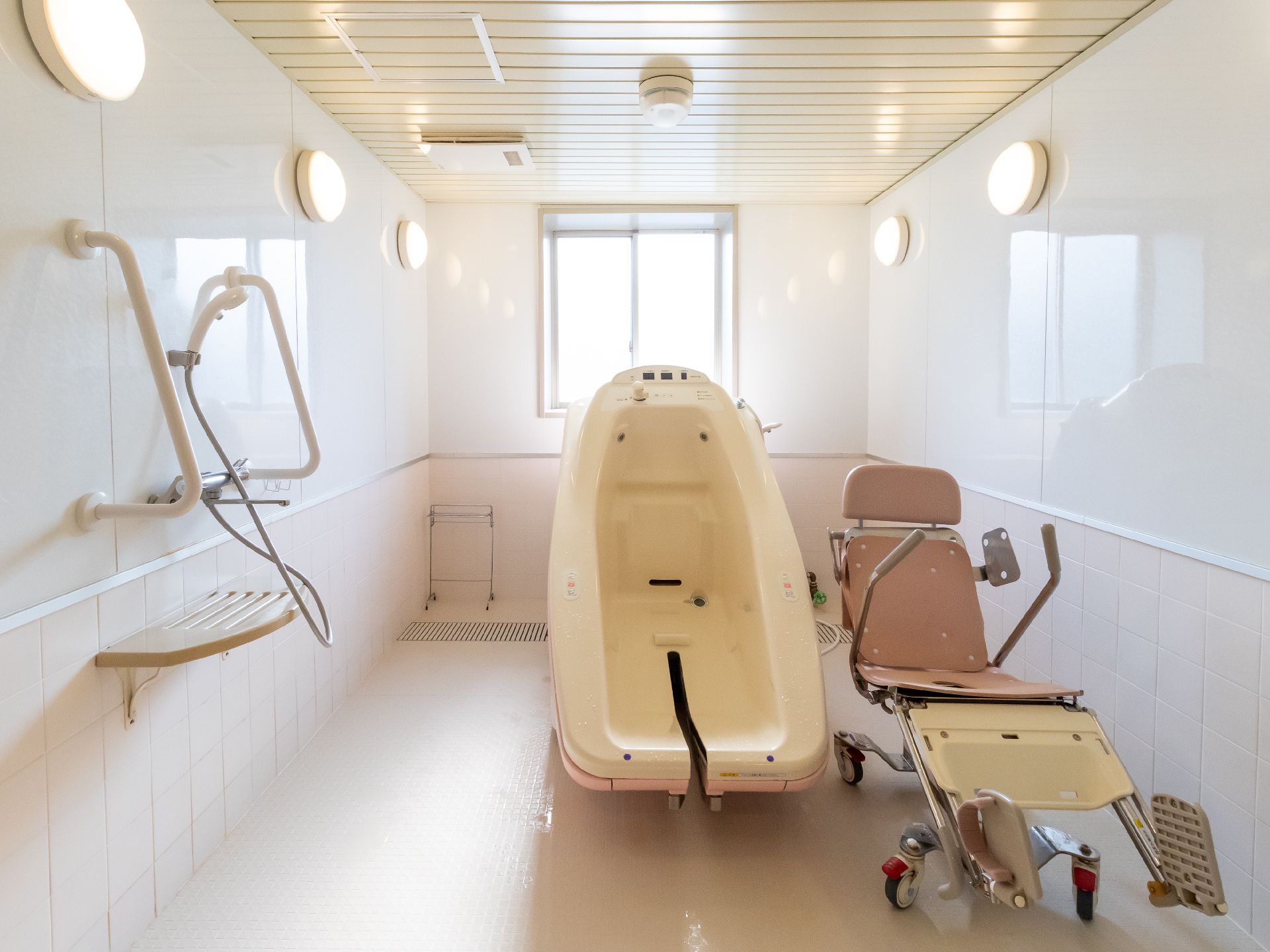 白い部屋に置かれたチェアー浴用の浴槽とピンクのシャワーキャリー