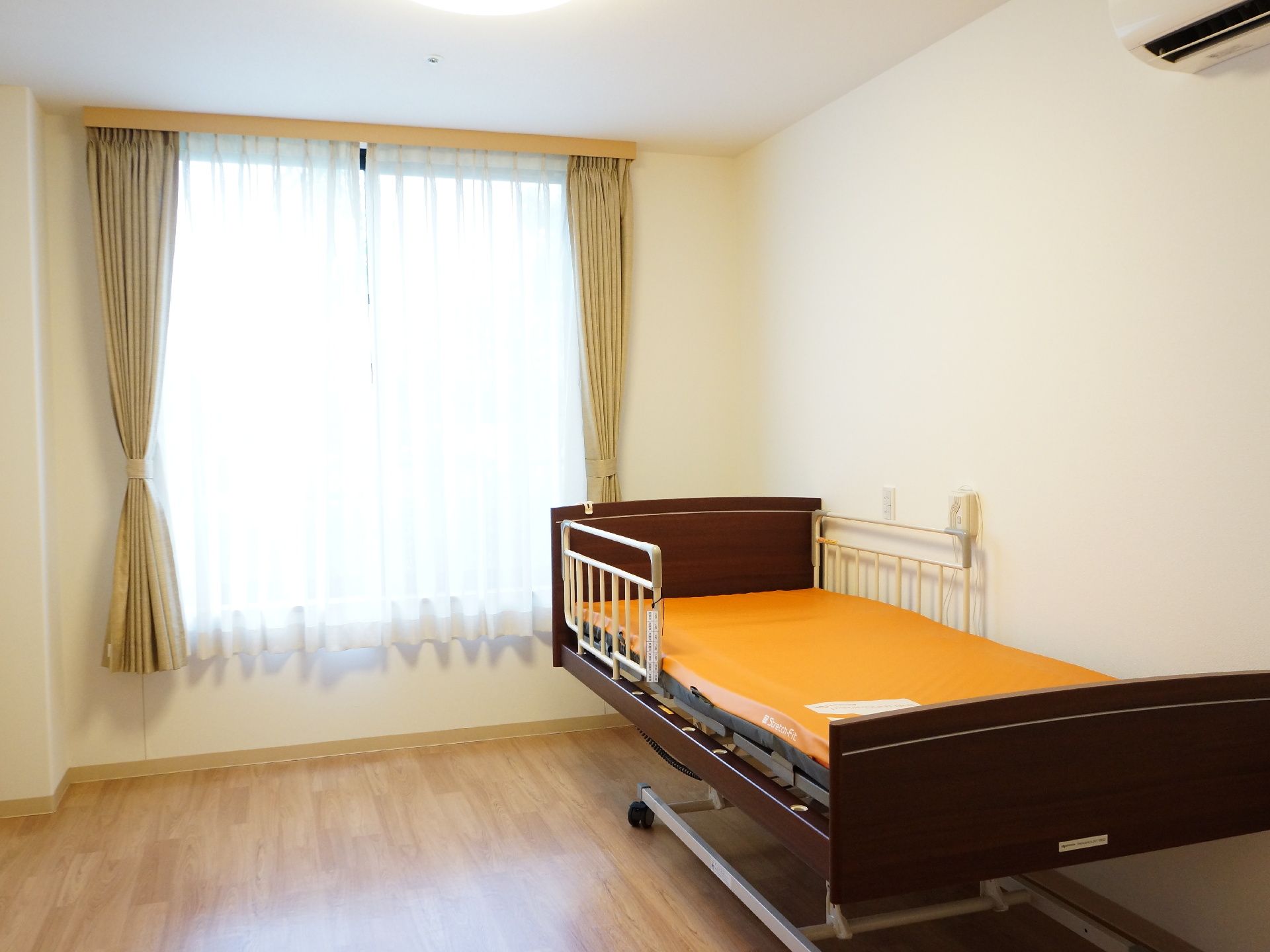 エアコンがついている。大きな窓があり、とても明るい部屋である。ベッドに手すりが付いている。車椅子の人も安全に利用できる。