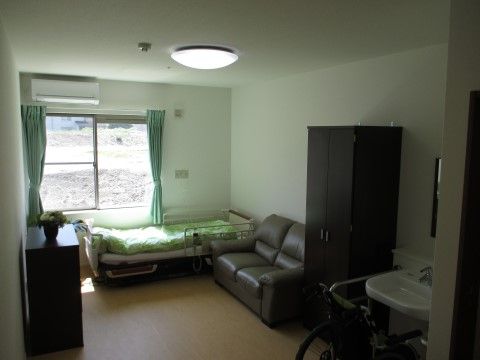室内にはエアコン、窓、洗面台が備えられている。フローリングの床の上には介護用のベッド、タンス、ソファ、ロッカーが置かれている。
