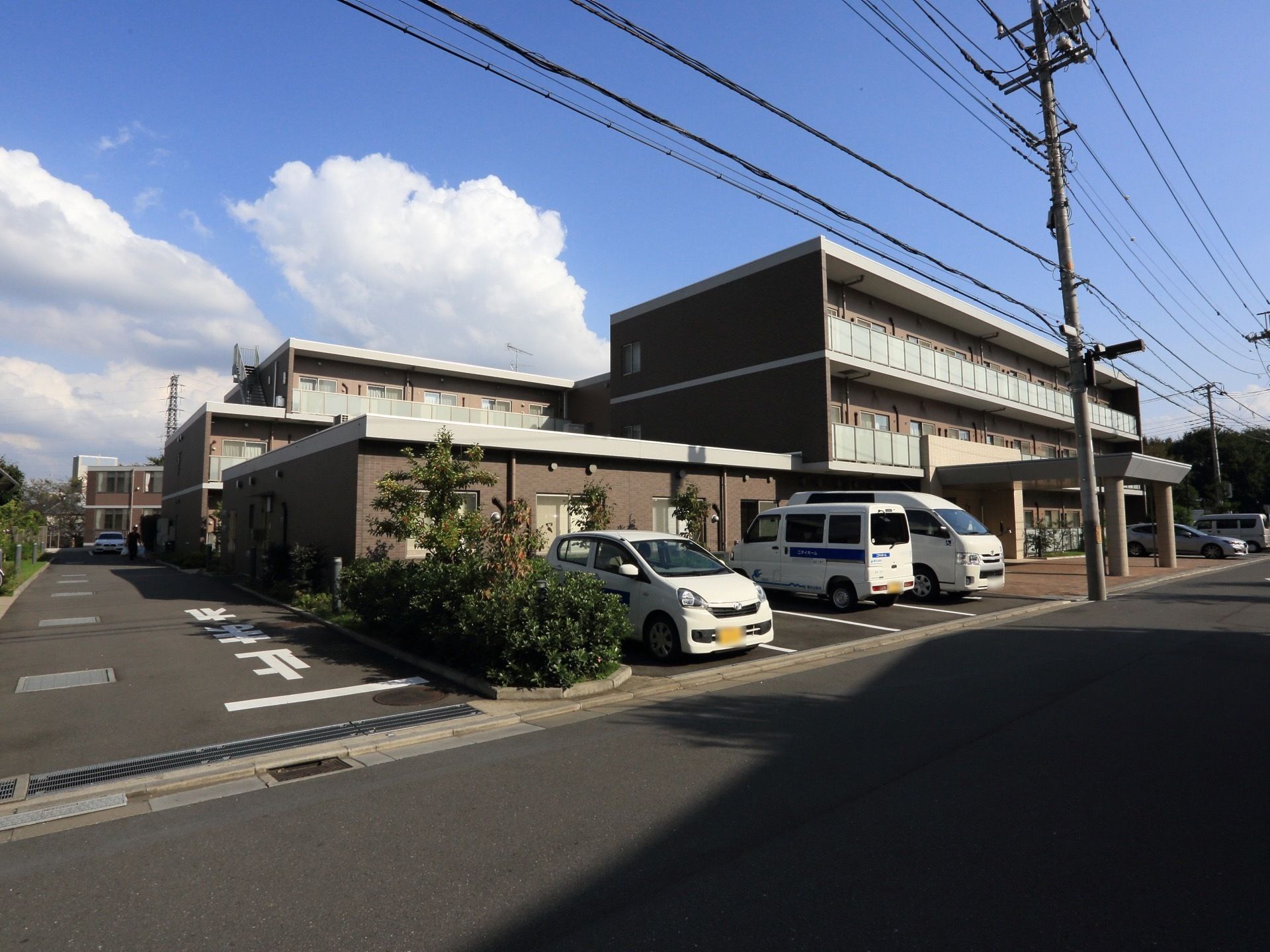 ３階建てのモダン雰囲気の外観である。駐車スペースが多いのが特徴。屋根が設置されているので雨の日でも濡れることなく室内に入れる。