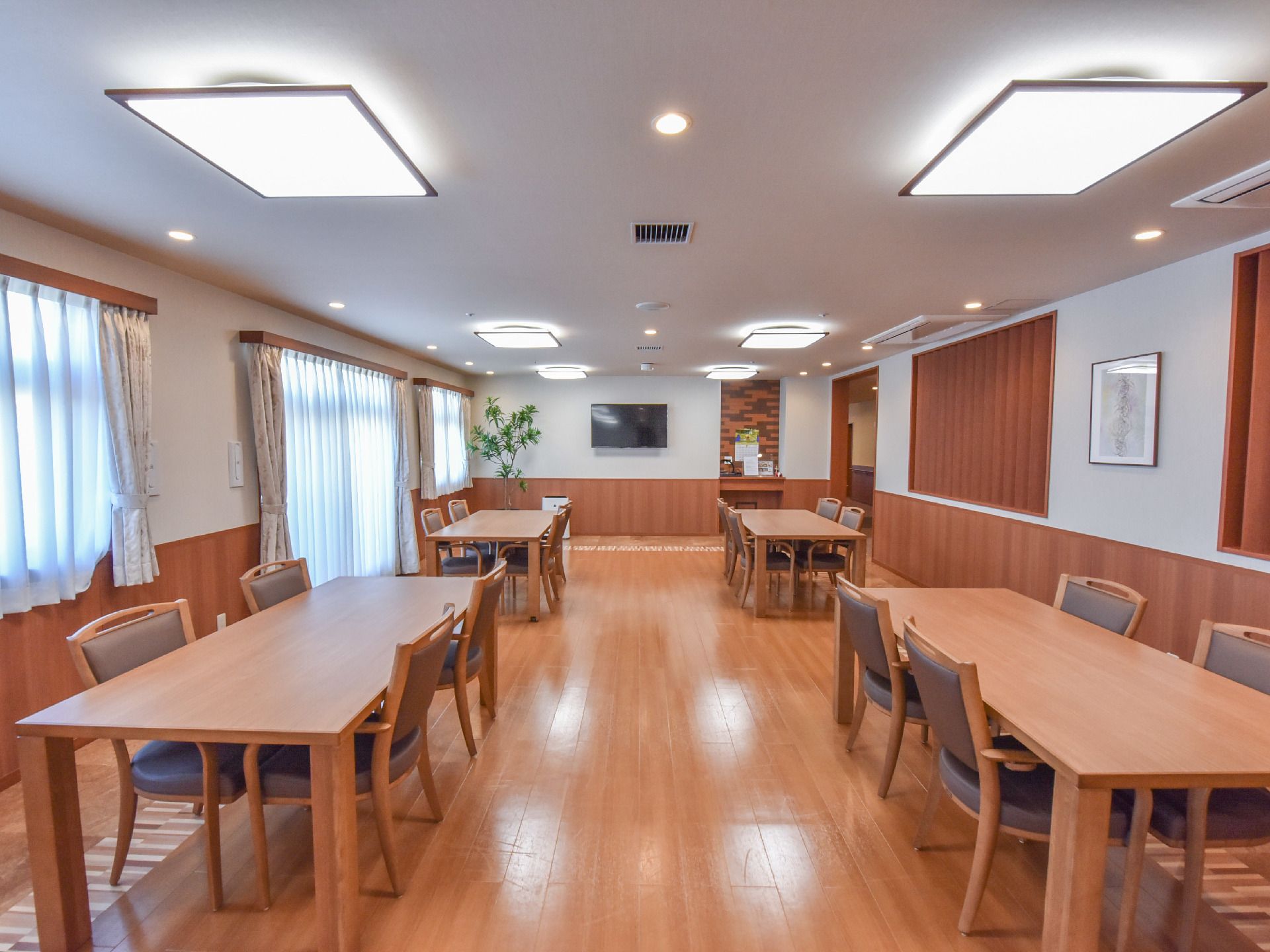 白色と木目の壁の部屋には大きい長方形のテーブルが4台ある。各テーブルには4脚の椅子が並べられている。