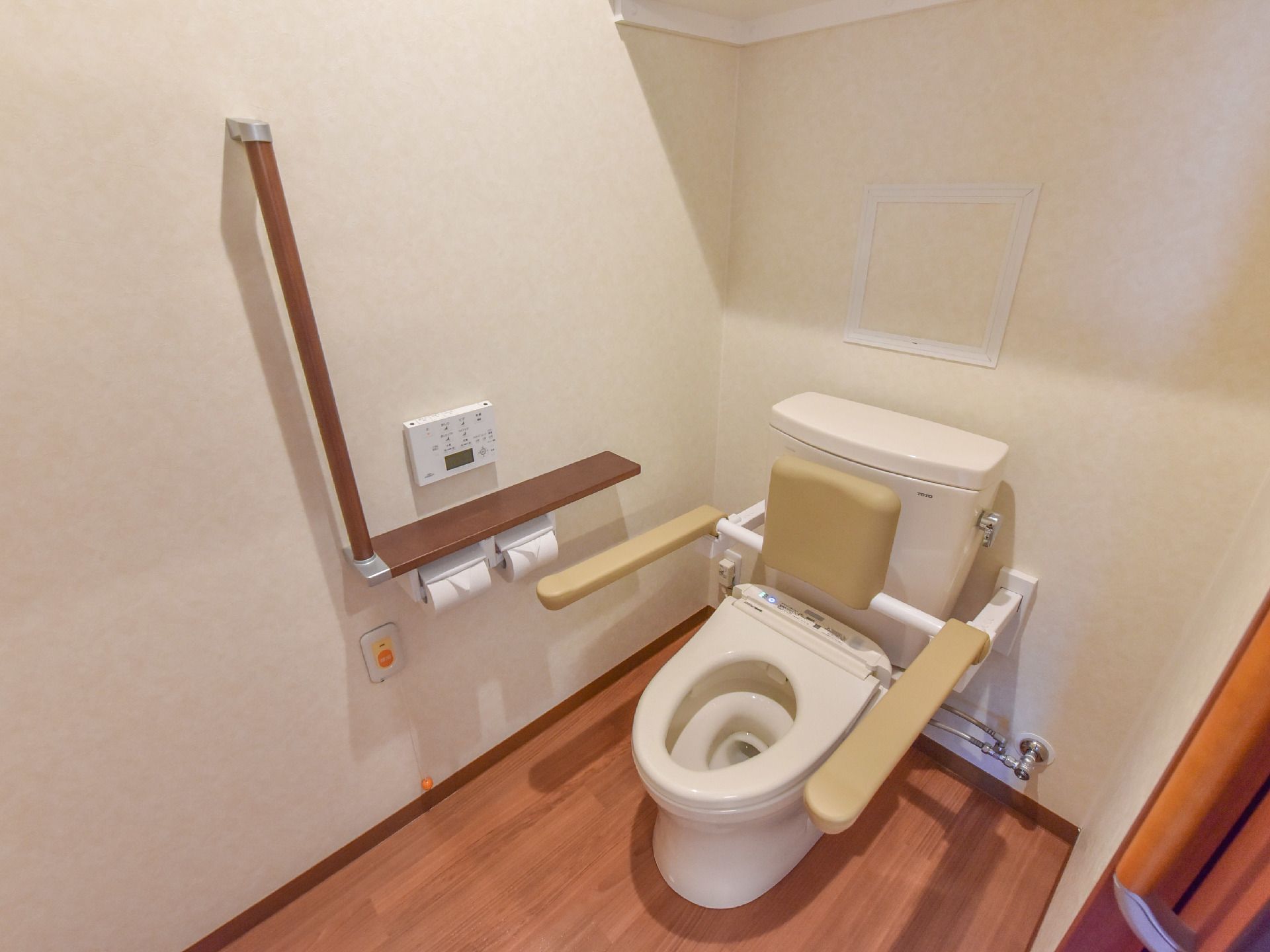 トイレには、ベージュ色の手すりと背もたれが取り付けられている。トイレットペーパーが2個あり、オレンジ色の呼出ボタンもある。