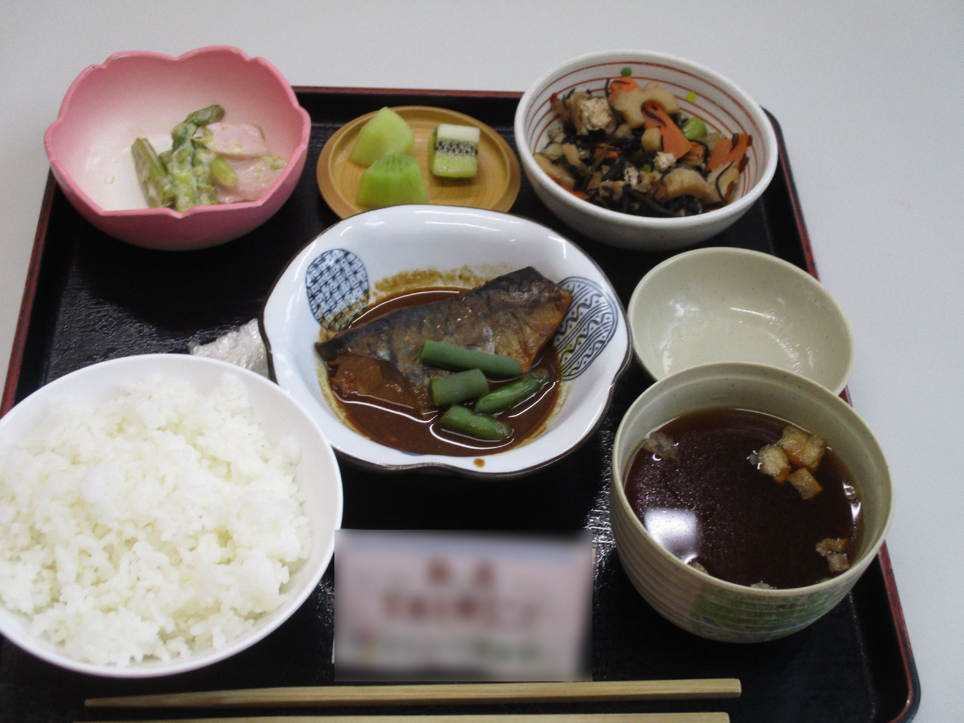 中央のさらには魚の煮込みがあり、他にもひじきの小鉢やフルーツ、ご飯にお吸い物が黒いトレイに乗せられている。
