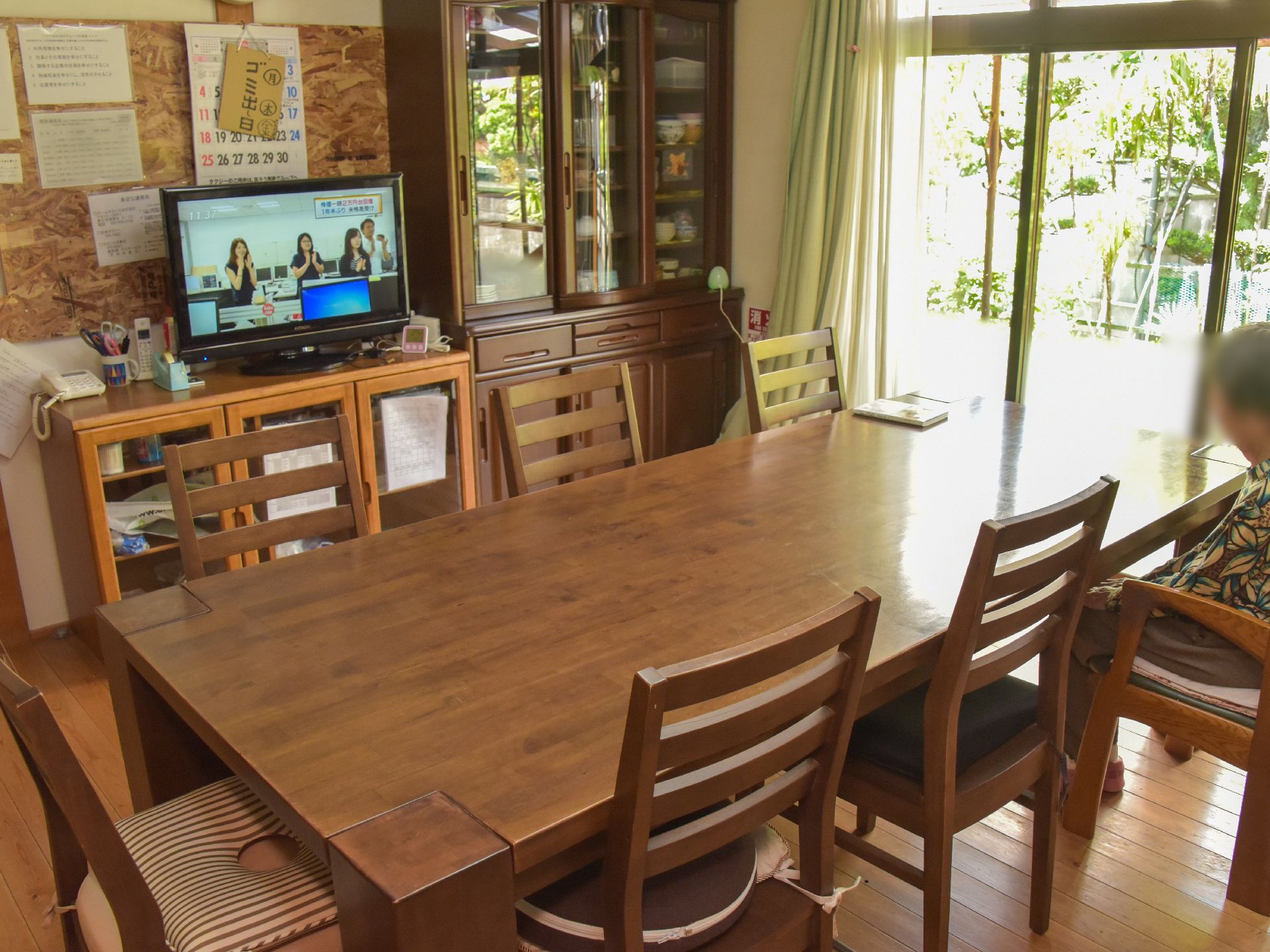 長机1台と椅子が7脚置いてある。テレビや食器棚があり、くつろぐことができる。大きな窓からは庭の風景が楽しめる。