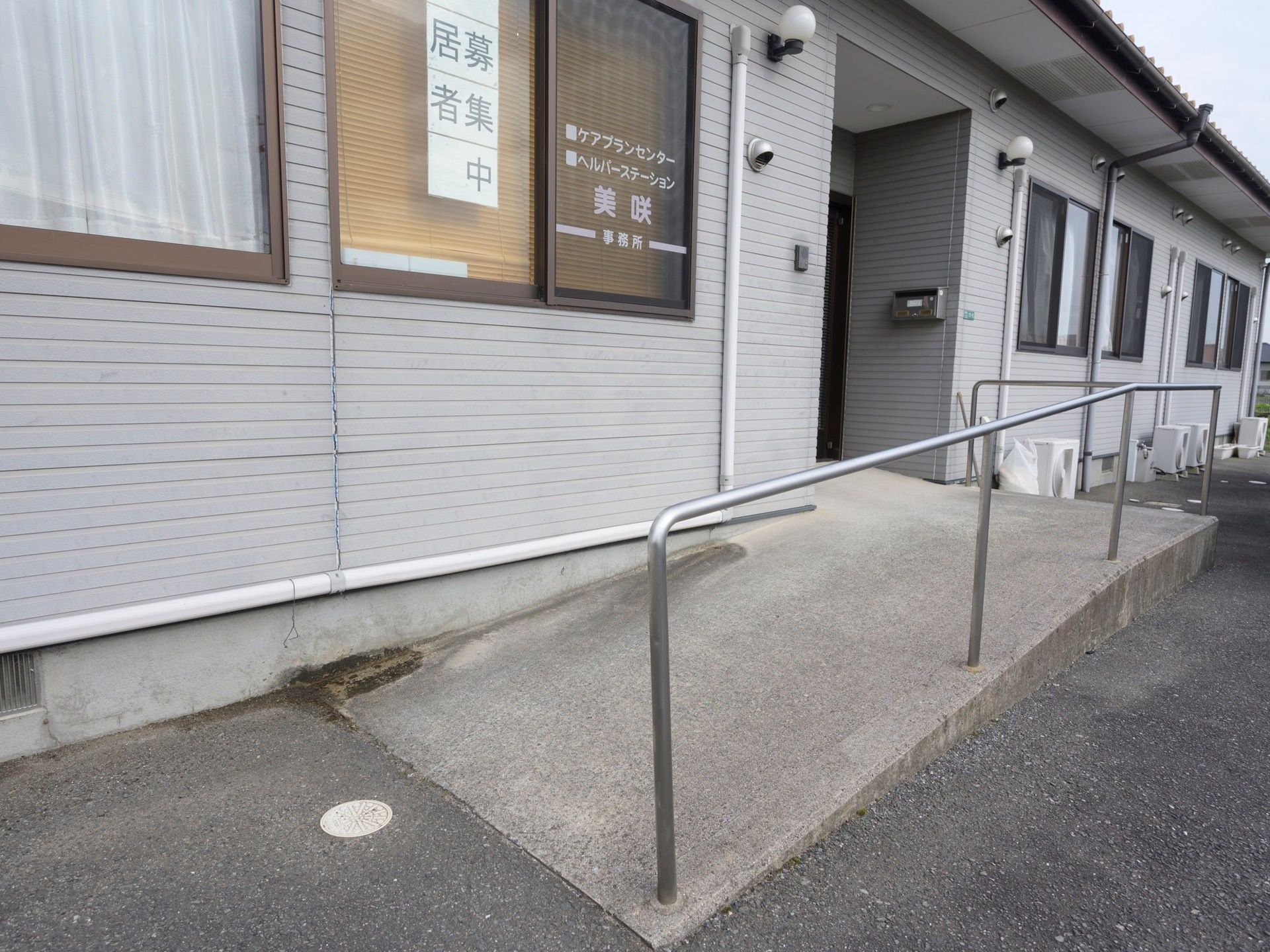 平屋建ての白い建物があり、玄関の前は緩やかなスロープになっている。スロープには手すりがある。
