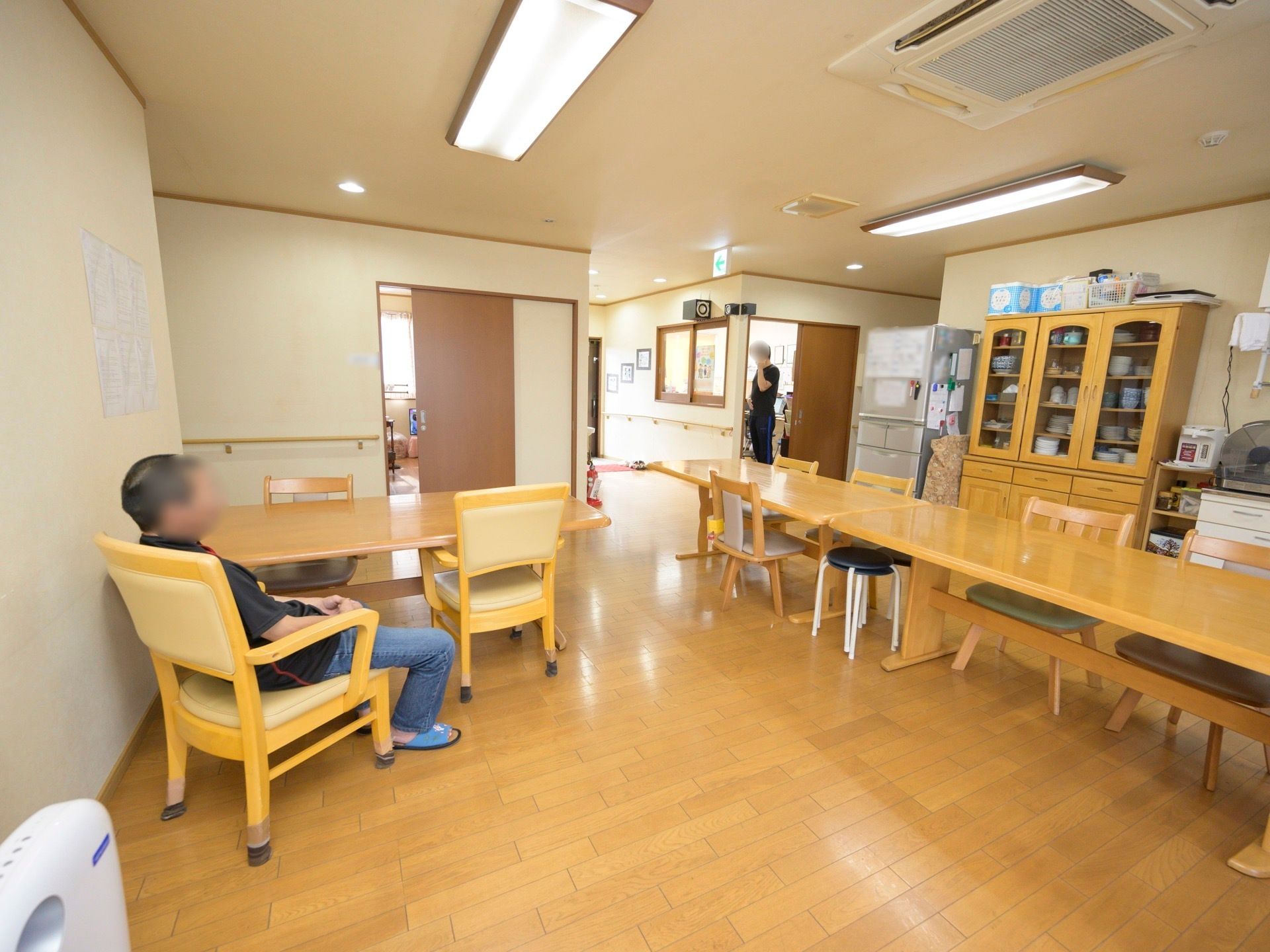 天井に空調の付いた広いフロアがある。ダイニングテーブルが並べられ、入居者様が１人席についてくつろいでいる。