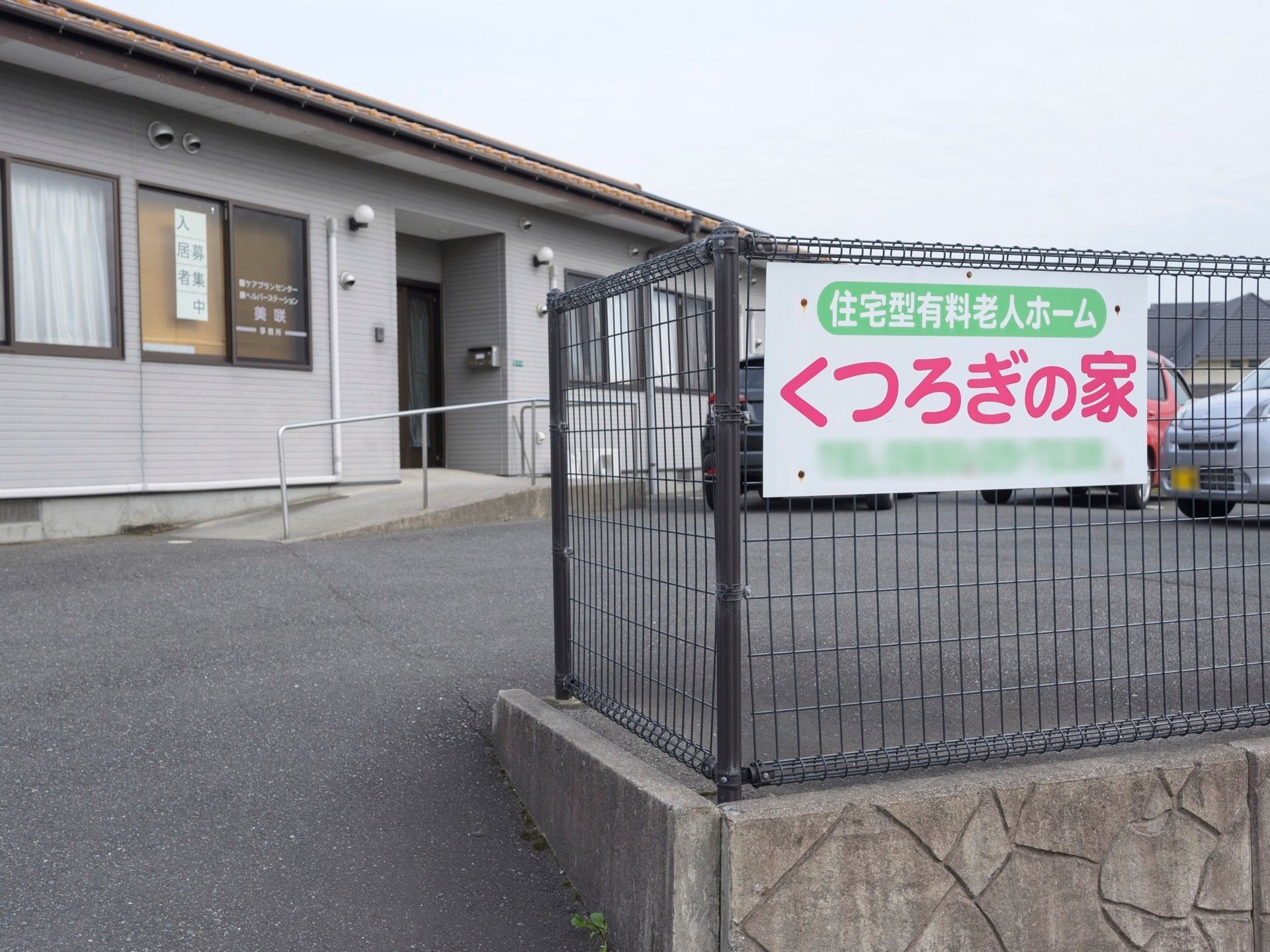アスファルトで舗装された敷地に、平屋建ての住宅が建っている。建物の前は駐車スペースになっており、車が数台停まっている。