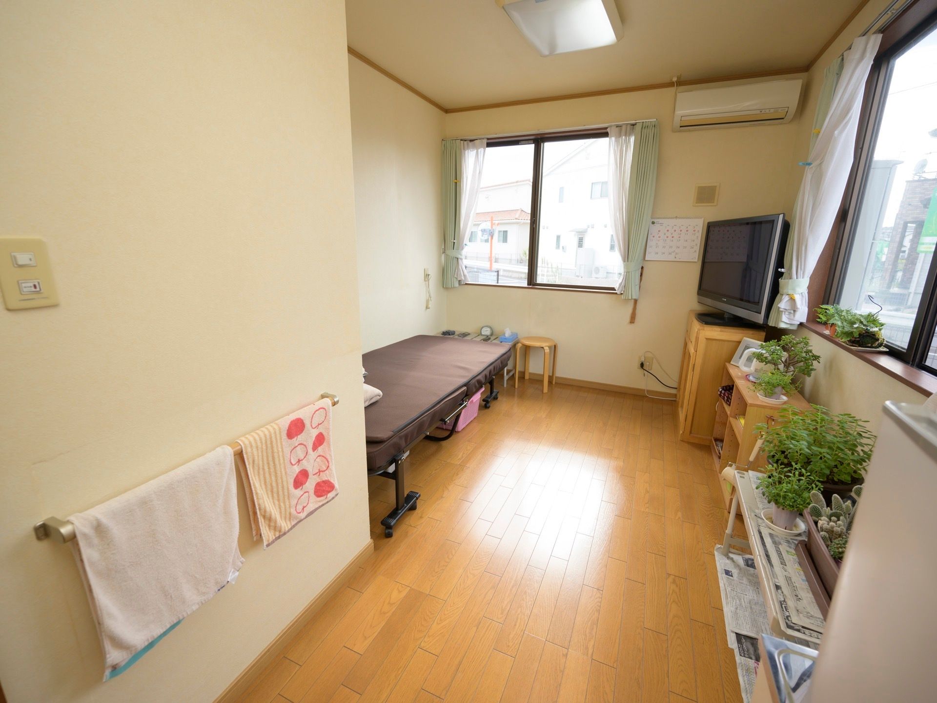 ２面に窓がある開放的な居室。壁際にベッドがあり、部屋の角にはエアコンと大型のテレビが設置されている。