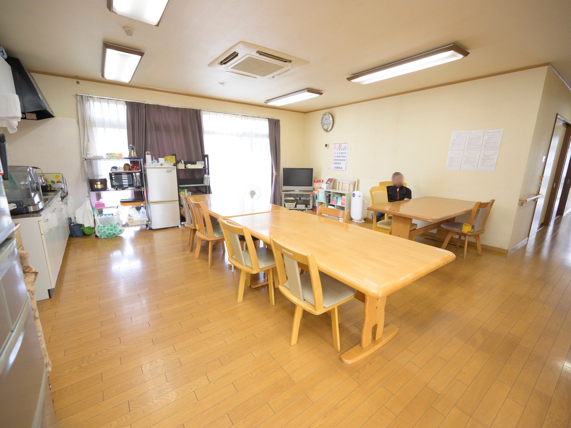 掃き出し窓がある明るい食堂。空調がついており、テーブルでは男性の入居者様が１人座ってくつろいでいる。
