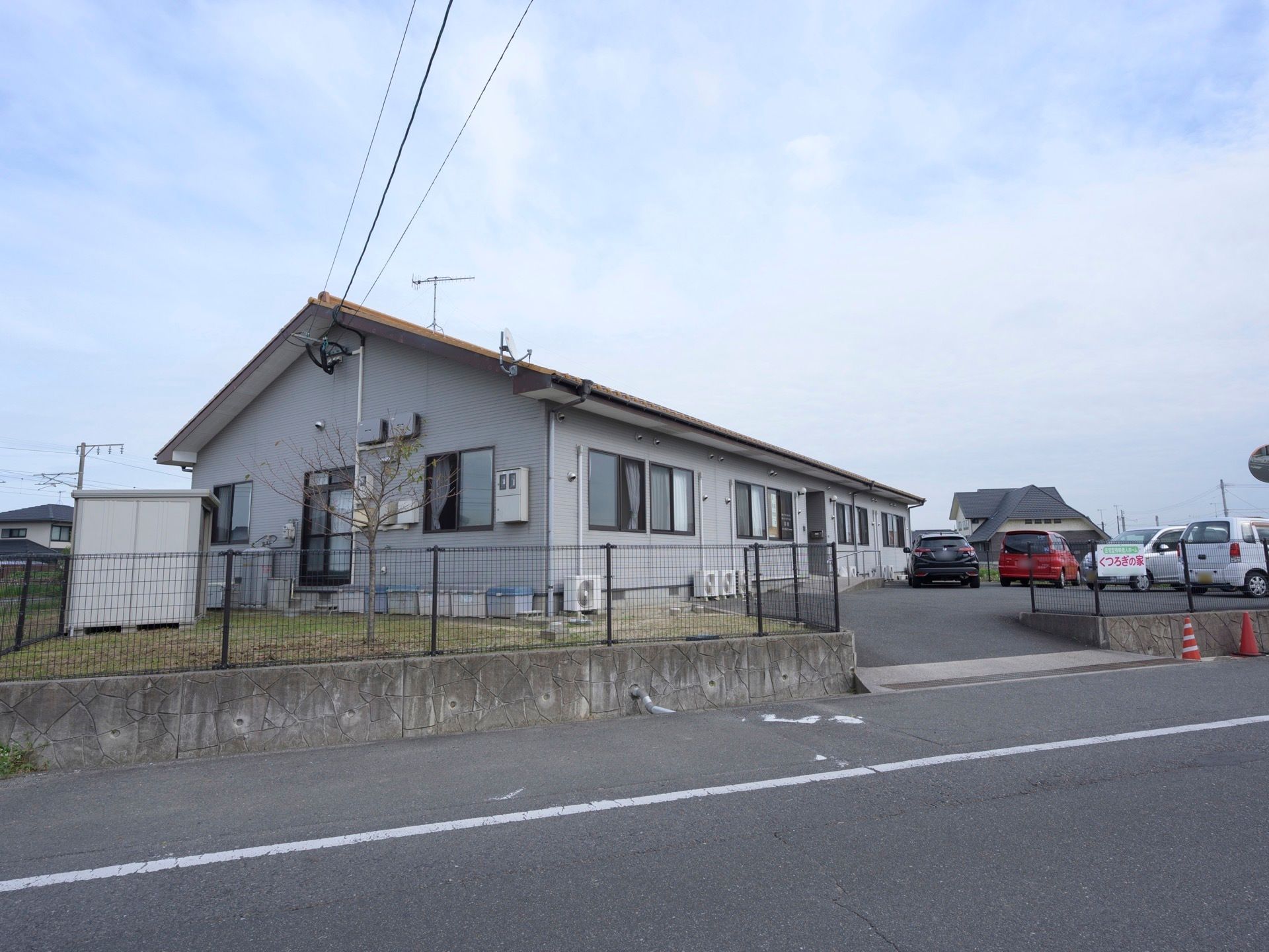 車道から少し奥まった場所に、平屋建ての住宅が建っている。敷地はフェンスで囲まれ、駐車場には車が数台停まっている。