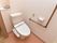 白色と暖かい味のある床のトイレスペースである。便器の横にはエル型の手すりがある。後方の斜め後ろの上には棚がある。