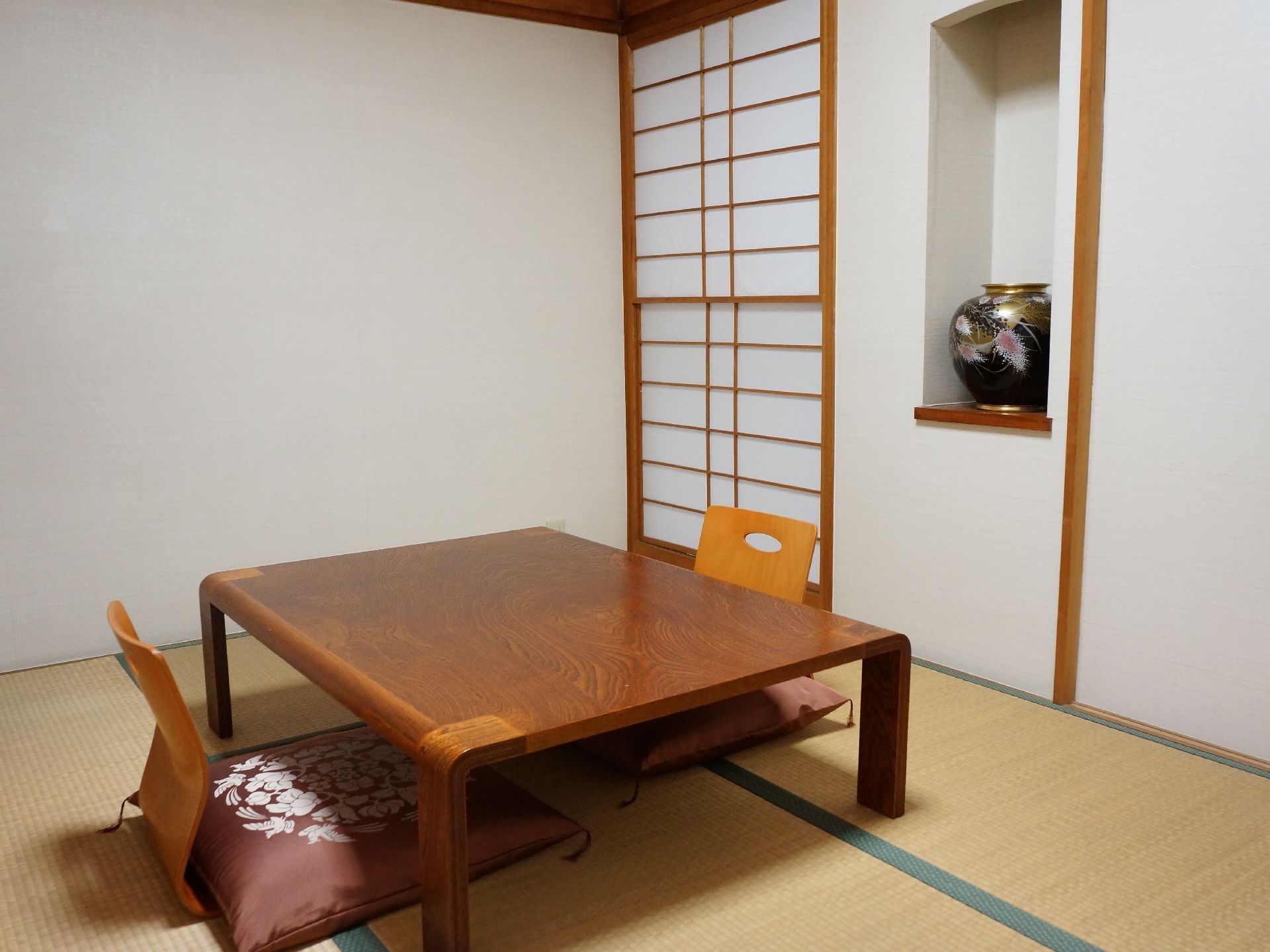 和室の部屋に、木製のテーブルがあり、座椅子2台に座布団が置かれている。部屋の隅に障子があり、その横にある棚に、花瓶が飾られている。