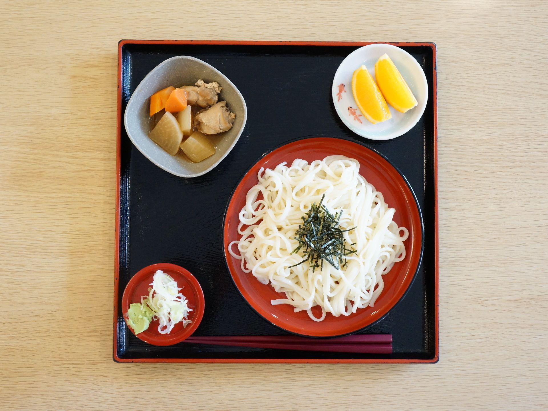 施設で提供される、食事の一例。黒いお盆の上に、メインの麺類がある。また、野菜の煮物、薬味、果物もある。