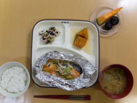左側にはご飯が一杯ある。右側には味噌汁がある。中央にあるプレートの中には魚の料理と副菜が二品添えられている。右上に果物がある。