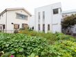 奥にはすずらんの建物がある。右奥には玄関がある。手前には様々な植物が生えている。周辺には民家が建っている。