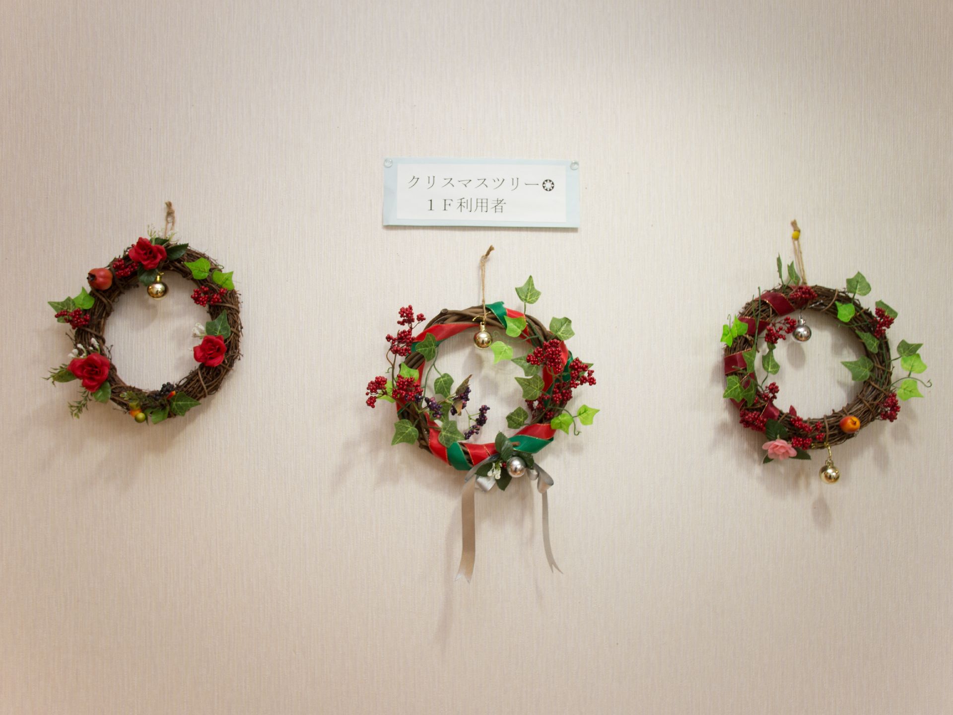 白地の壁がある。壁には三つのリースが飾られている。リースの上にはクリスマスツリー1F利用者と書かれた紙が貼りつけられている。