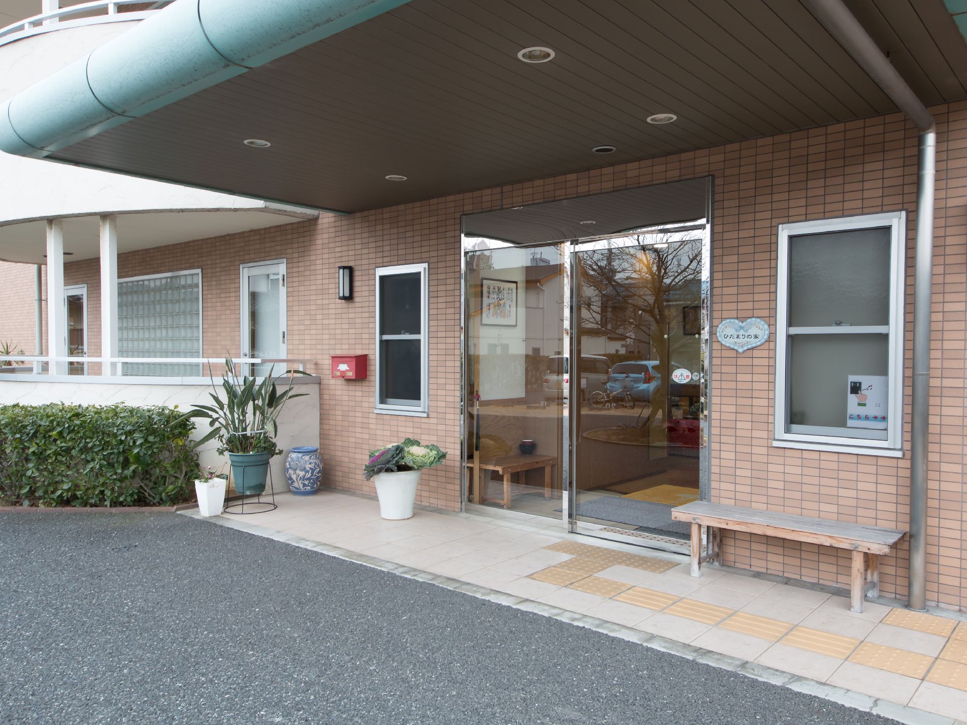 玄関入口には大きな屋根が設置されている。自動扉の前には植物、横側にはベンチが置かれている。床には点字ブロックがある。