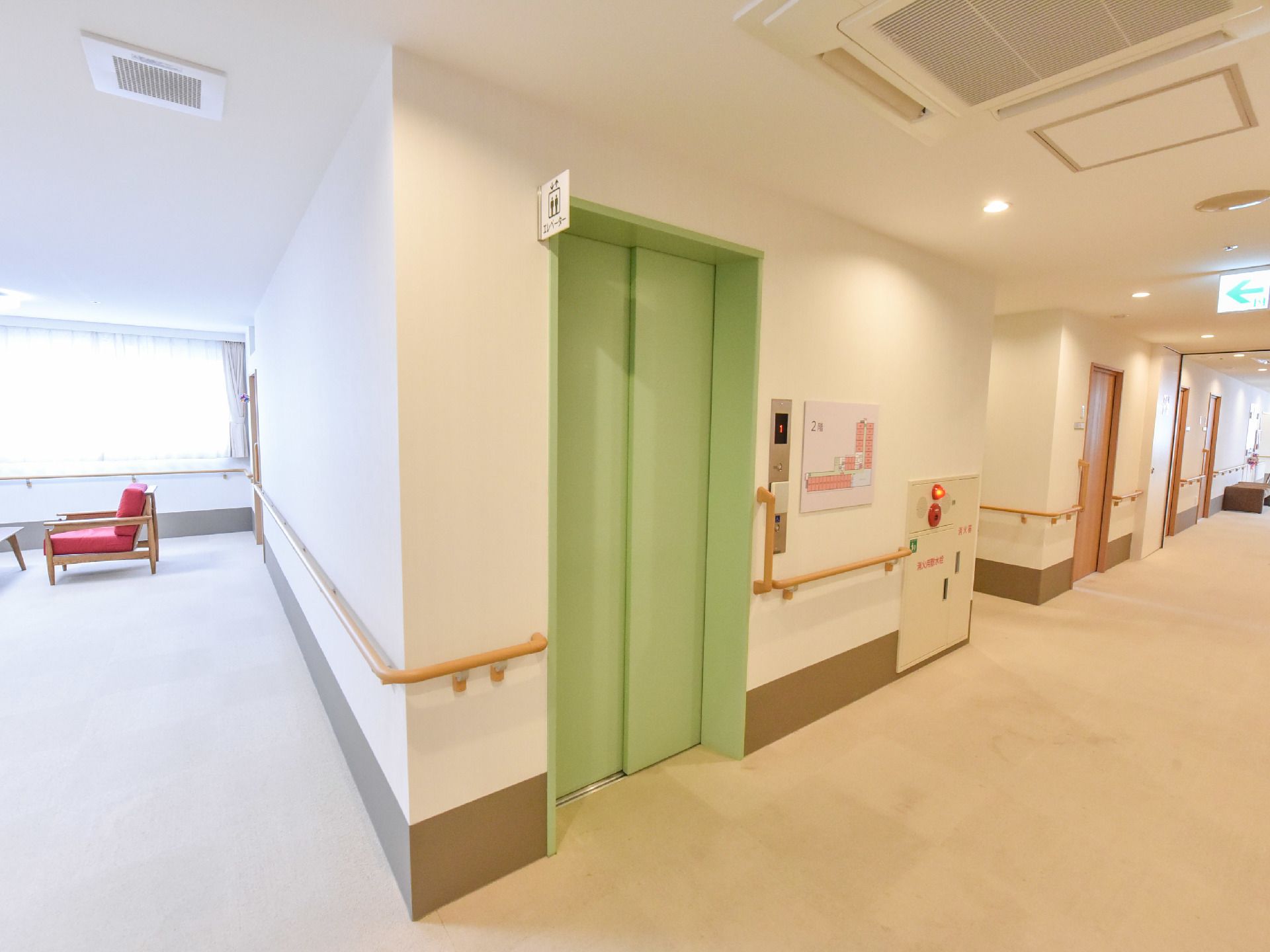 椅子とテーブルが置かれたフロアの近くにはエレベーターが1機ある。エレベーター横には病院案内の地図と手すりが設置されている。