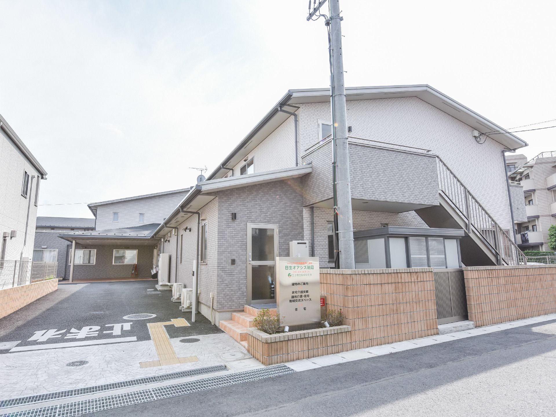 施設は道路に隣接している。道路側にある植え込みには施設の看板が取り付けられている。施設の外階段も見える。