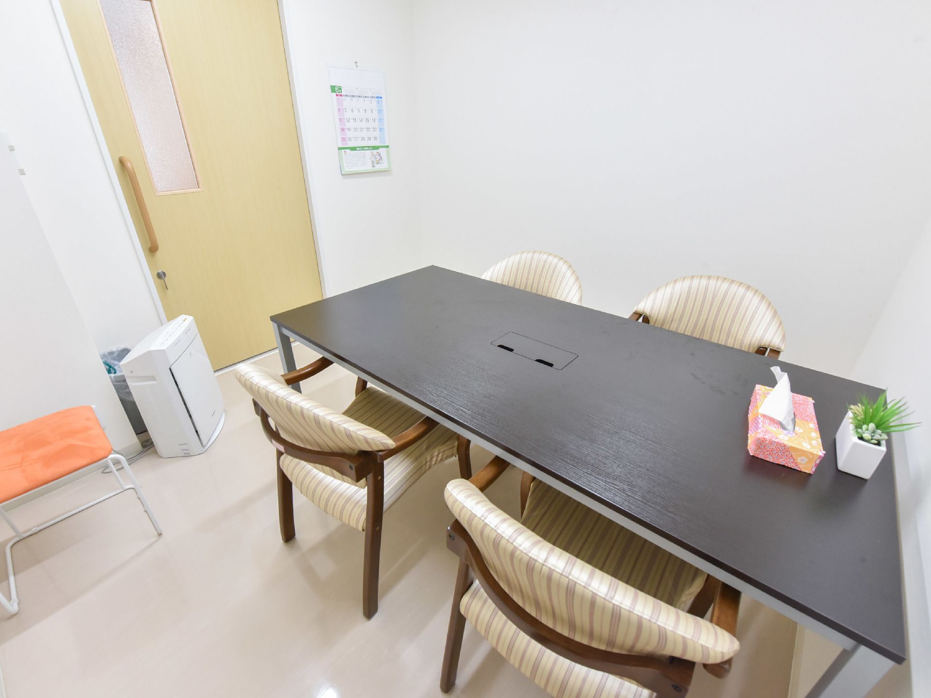 カレンダーが壁に付けられた部屋にはテーブルと椅子が置かれている。テーブルにはミニ植物とテッシュボックスがある。