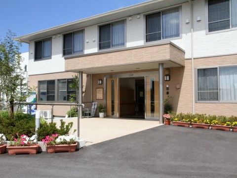 2階建て施設の玄関には、両開きの自動扉が設置されている。入口ポーチが広く確保され、手入れの行き届いた花々が配置されている。