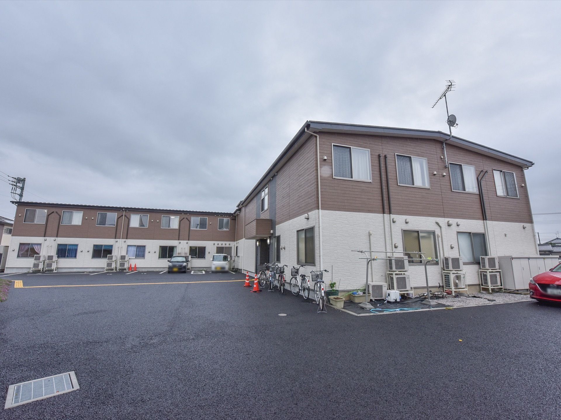 駐車スペースに自動車と自転車が複数停まっている。施設の外装は茶色と白で構成されている。室外機がいくつか設置されている。