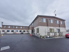駐車スペースに自動車と自転車が複数停まっている。施設の外装は茶色と白で構成されている。室外機がいくつか設置されている。