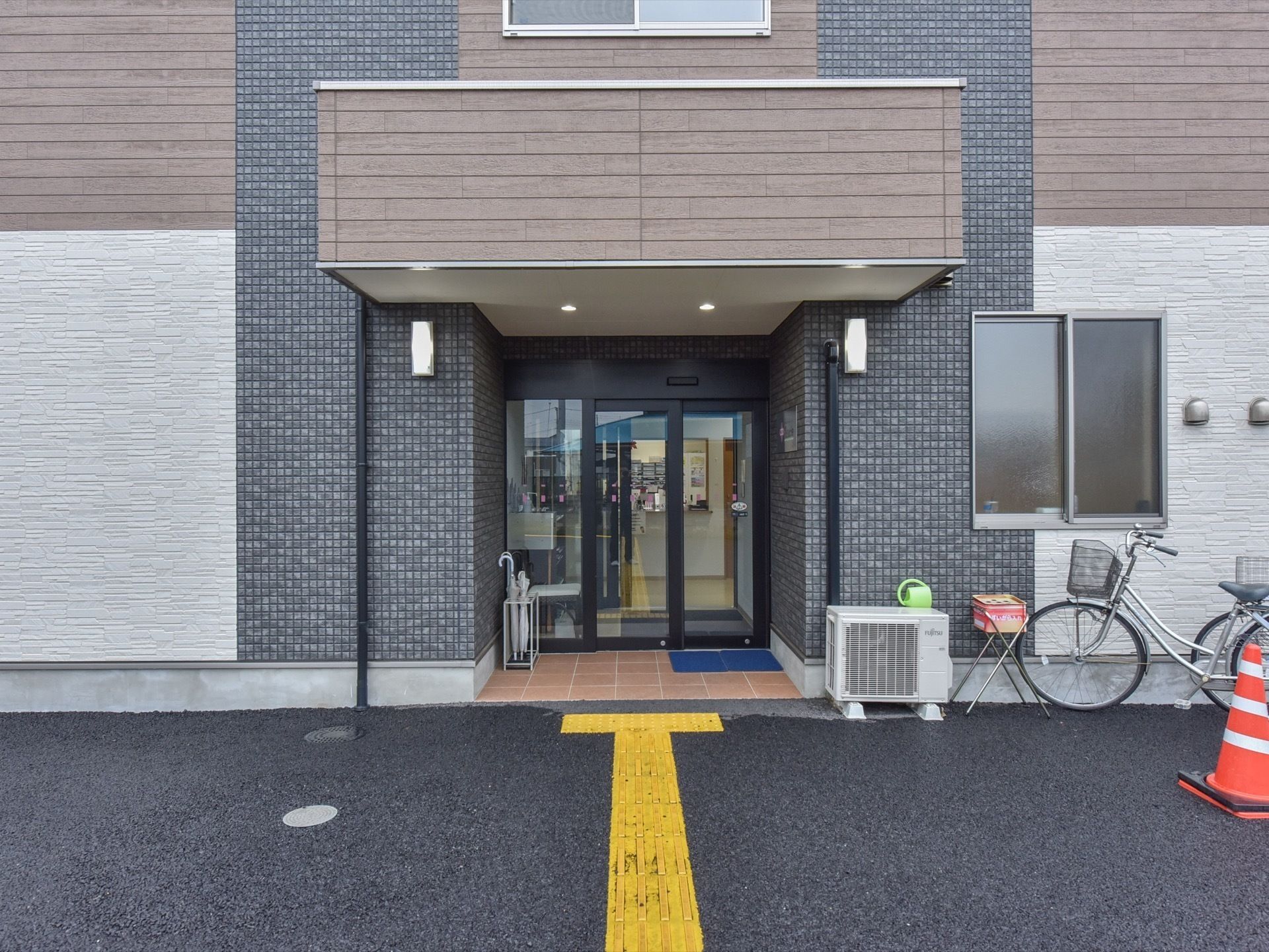 アスファルトが濡れている。玄関には黒いタイルが使われている。傘立てがある。自転車が１台停まっている。室外機がある。