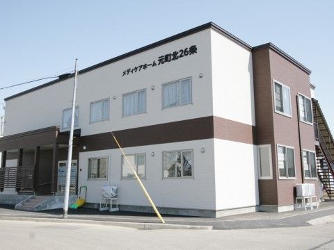 白色と茶色い外観の施設がある。黒い立体文字で施設名が書かれたプレートが貼られている。窓にはカーテンがかけられている。