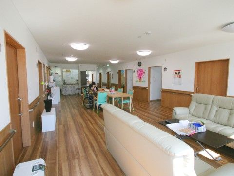 食堂は広々としている。テーブル席には緑色の椅子があり、車椅子に座った入居者様が座っている。離れた場所にはソファーが置かれている。