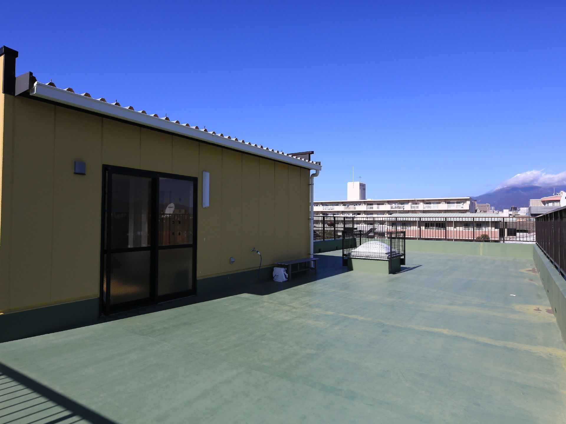 フェンスで囲まれた屋上で、緑色に床が塗られている。街並みの向こうには、大きな山が見えており雲がかかっている。