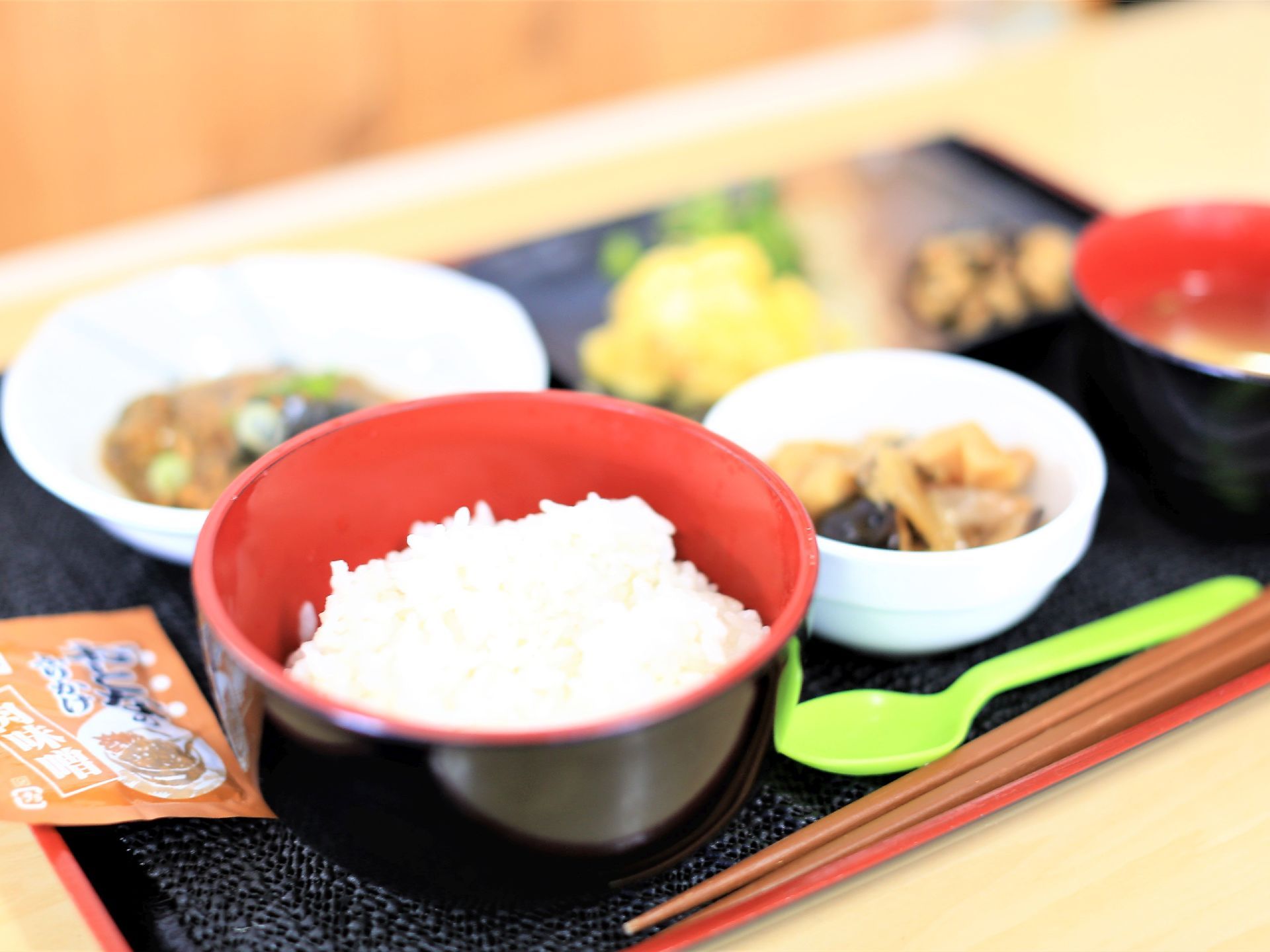 ご飯、味噌汁、煮物など彩り豊かな食事が盆の上に置かれている。ふりかけがついている。お箸とスプーンが用意されている。