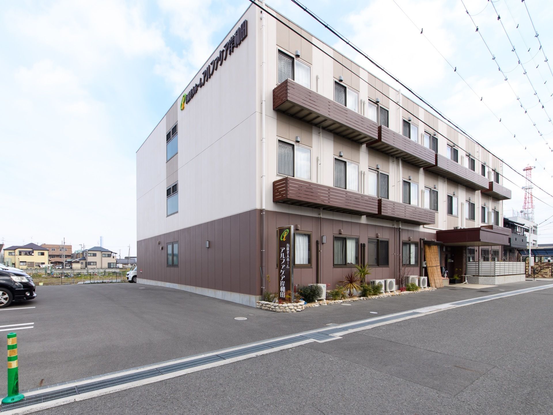 ベージュとブラウンの温かい雰囲気の外壁の3階建て。居室の窓には花台が設置されている。駐車場が隣接されている。